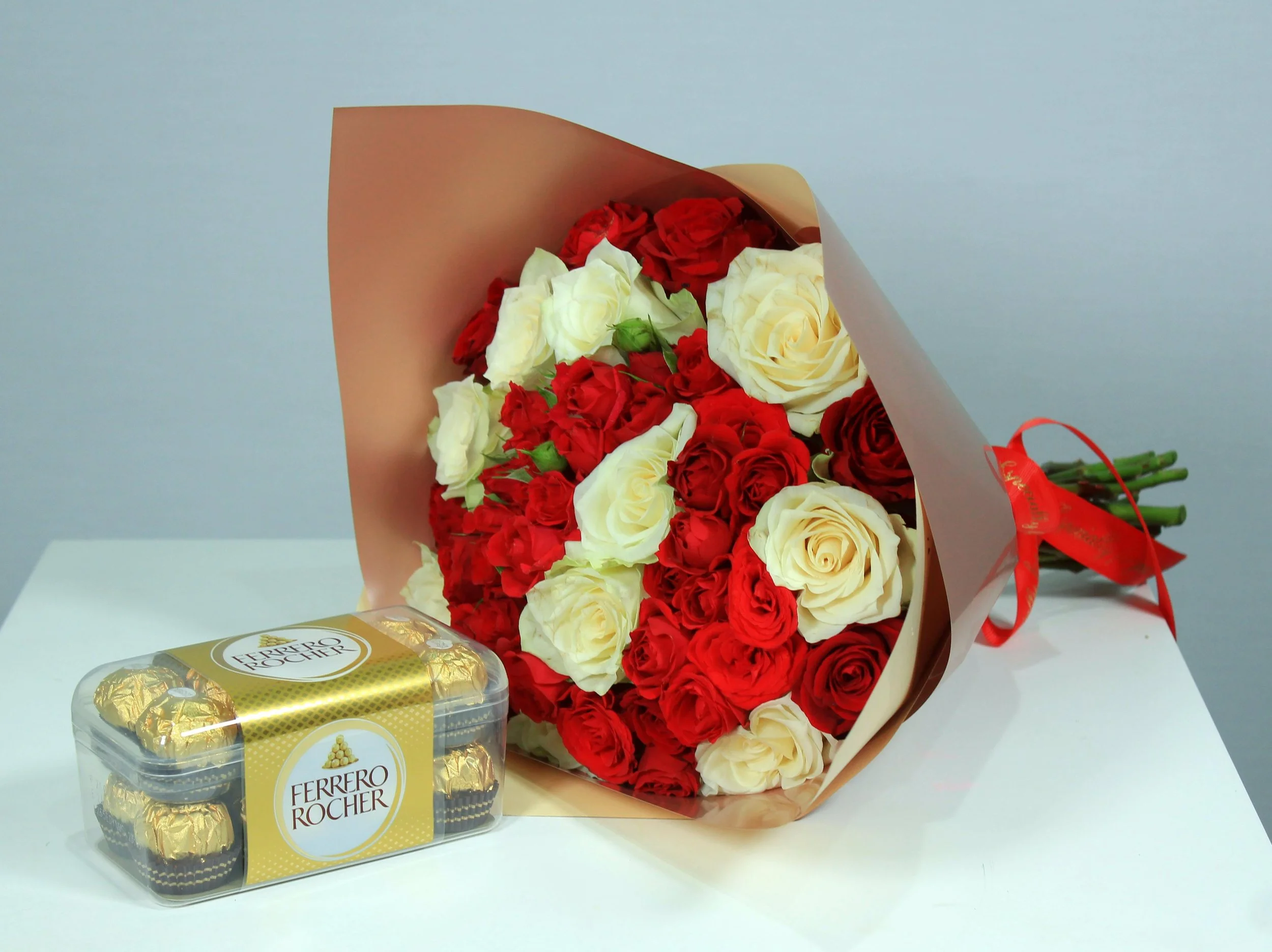 A bouquet of red and white roses wrapped in brown paper and tied with a red ribbon, placed on a white surface next to a box of Ferrero Rocher chocolates.