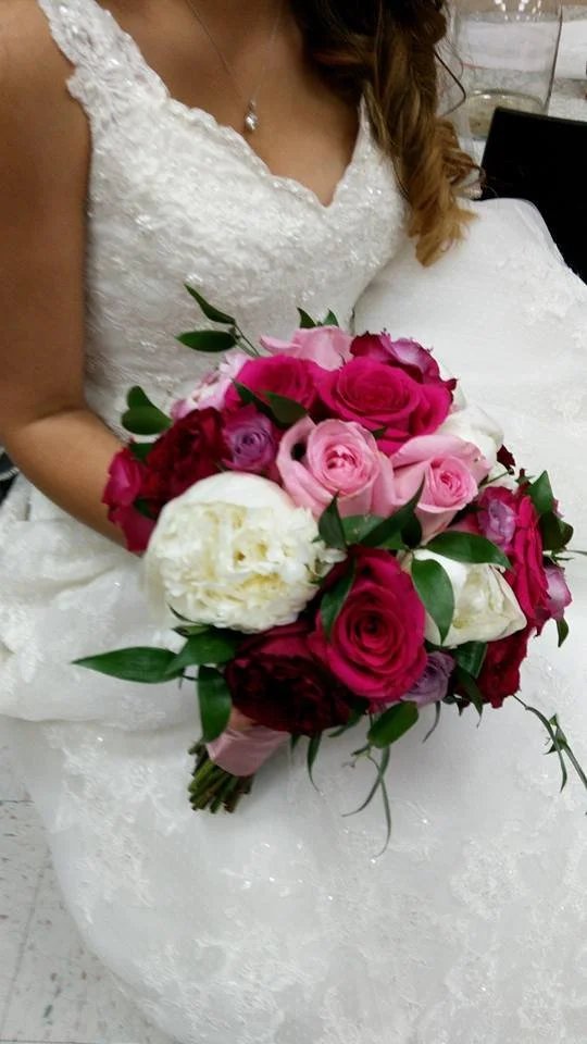 Bride in a white wedding gown holding a bouquet of pink, red, and white roses with green leaves.