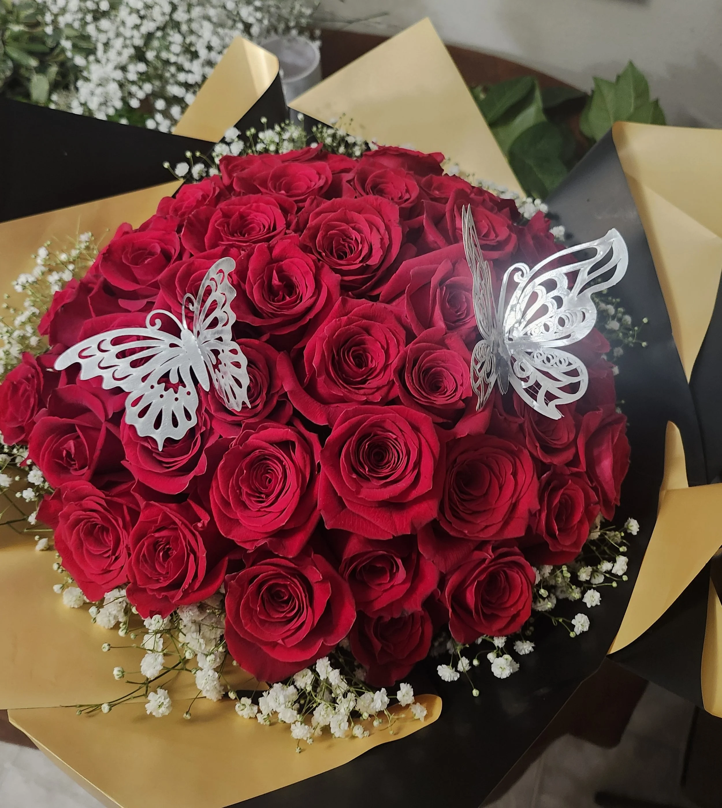 A bouquet of red roses with white baby's breath flowers, decorated with two silver butterfly ornaments, wrapped in black and gold paper.