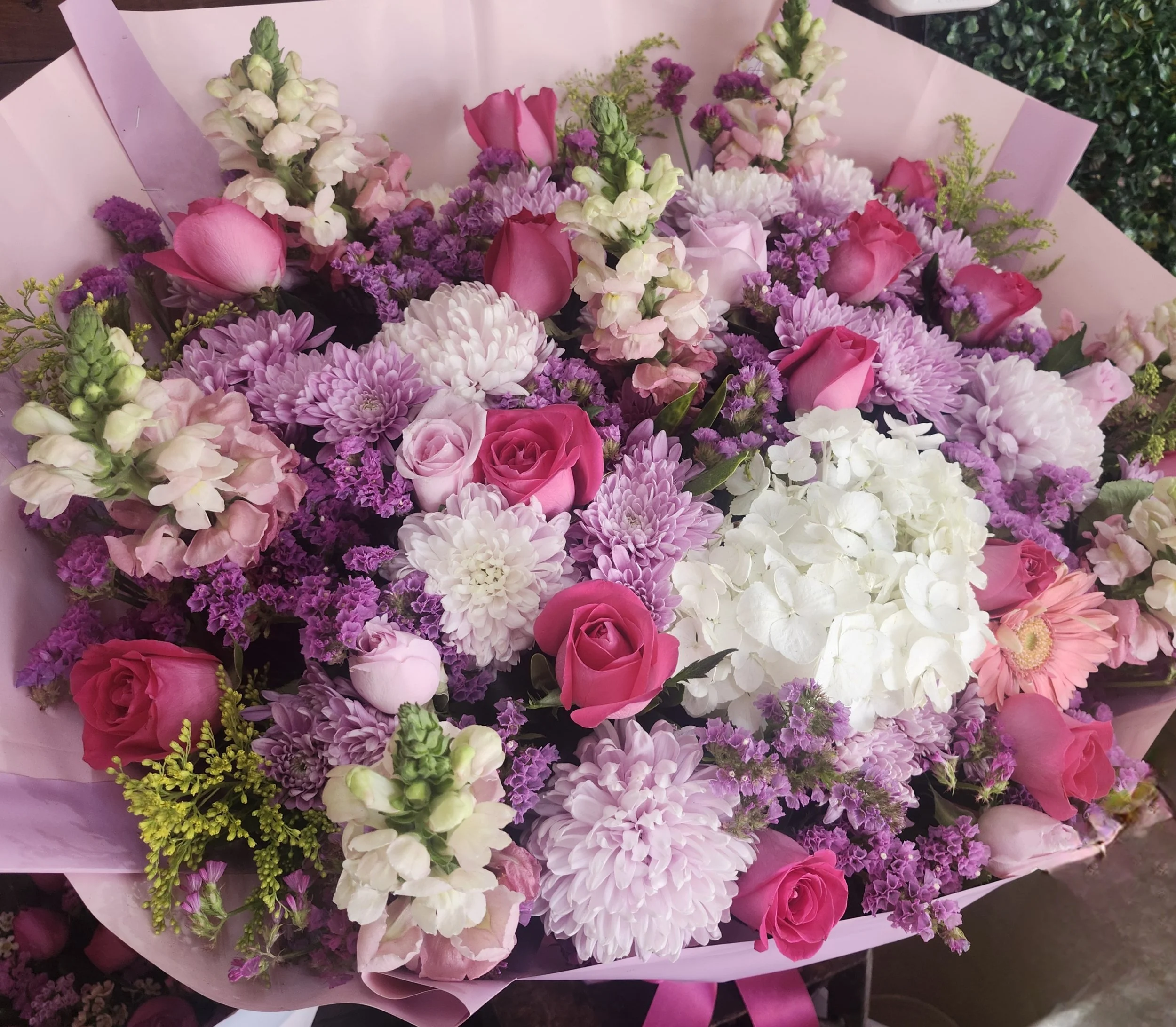 A bouquet of pink, purple, and white flowers including roses, dahlias, hydrangeas, and snapdragons wrapped in pink paper.