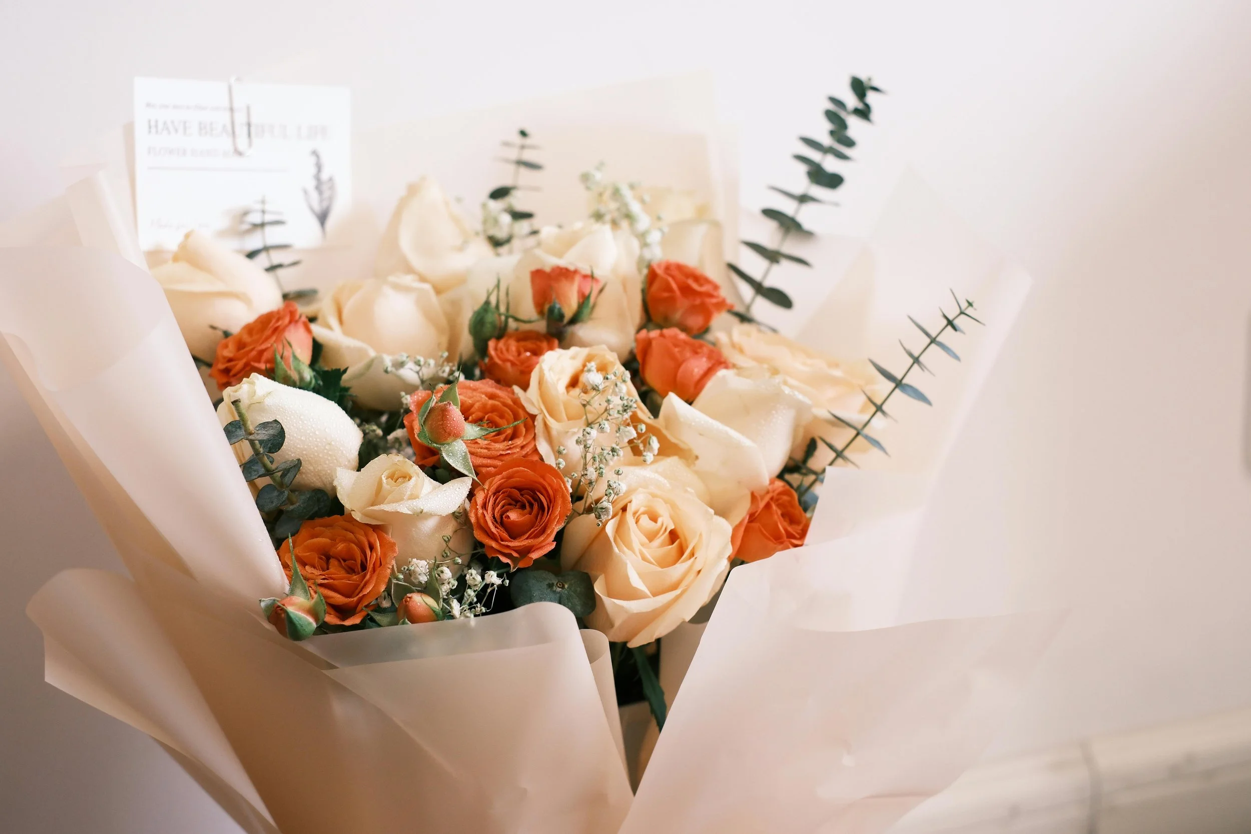 A bouquet of white and orange roses with greenery, wrapped in white paper.