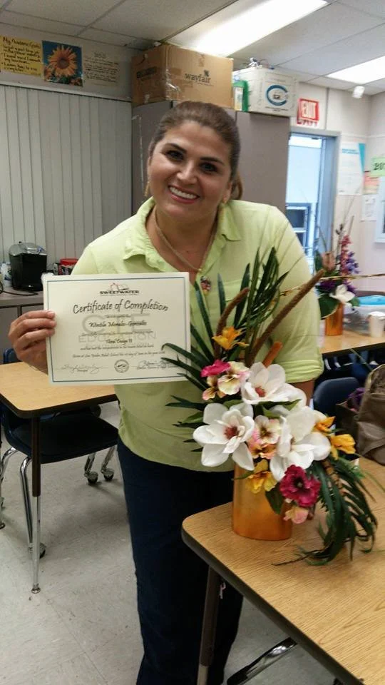 A woman smiling and holding a certificate of completion in a classroom, with a colorful floral arrangement on the table in front of her.