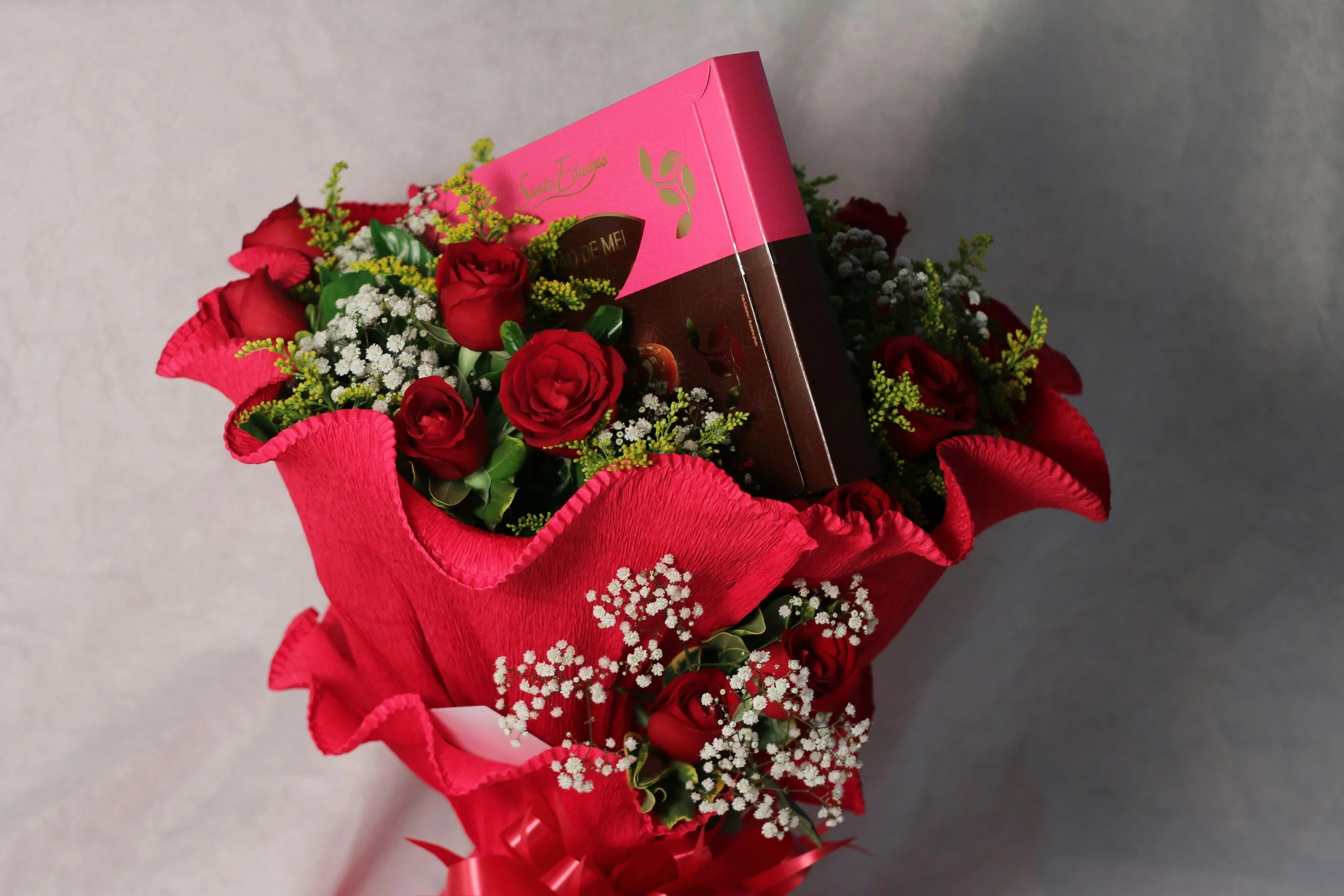 A bouquet of red roses, white baby's breath, and green foliage wrapped in pink and red decorative paper, with a pink box partially visible among the flowers.