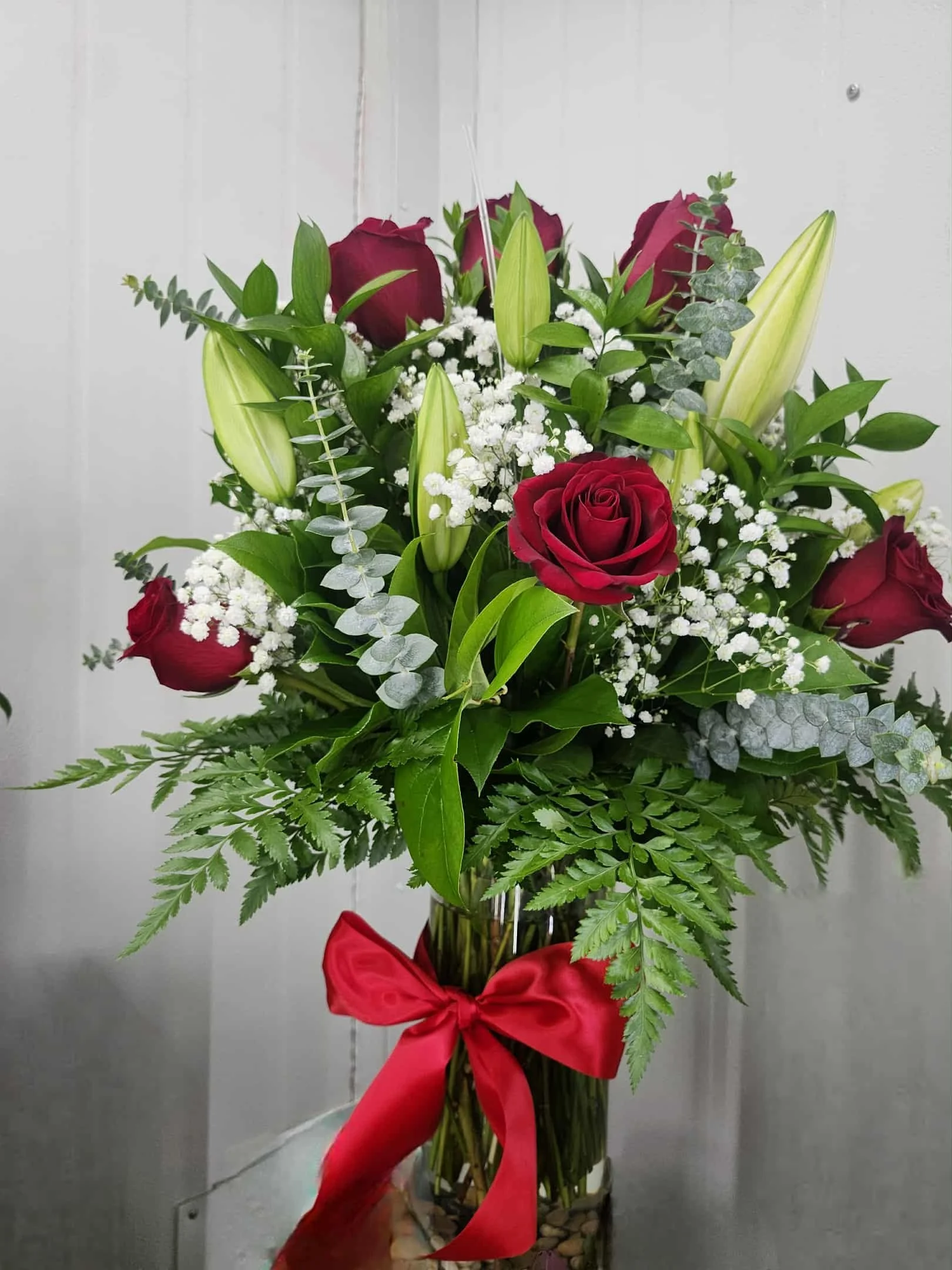 A bouquet of red roses, white lilies, baby's breath, and mixed green foliage in a glass vase with a red ribbon bow.