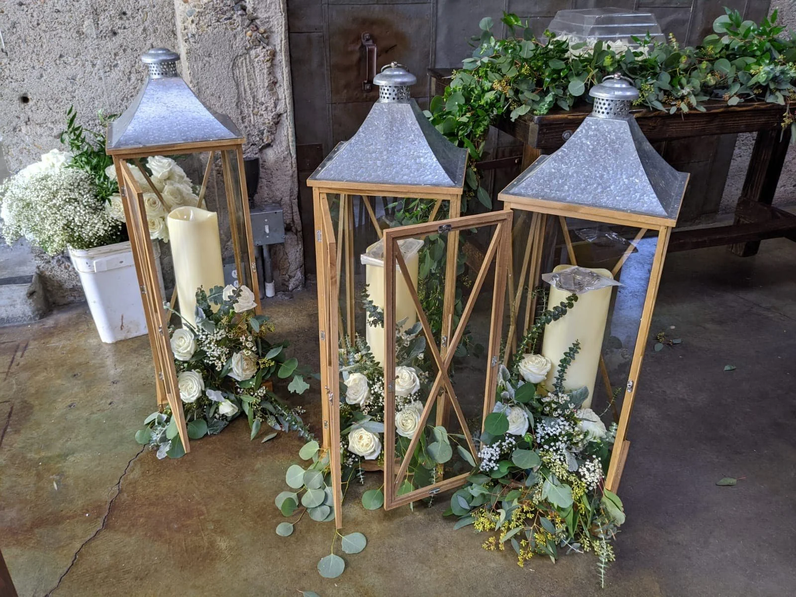 Three tall lanterns with candles and floral arrangements featuring white roses and greenery, placed on a concrete floor outdoors.