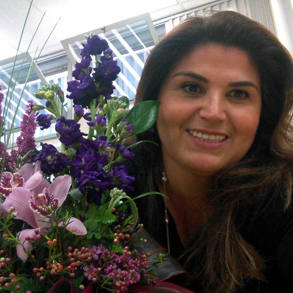 A woman with long brown hair smiling next to a bouquet of purple and pink flowers.