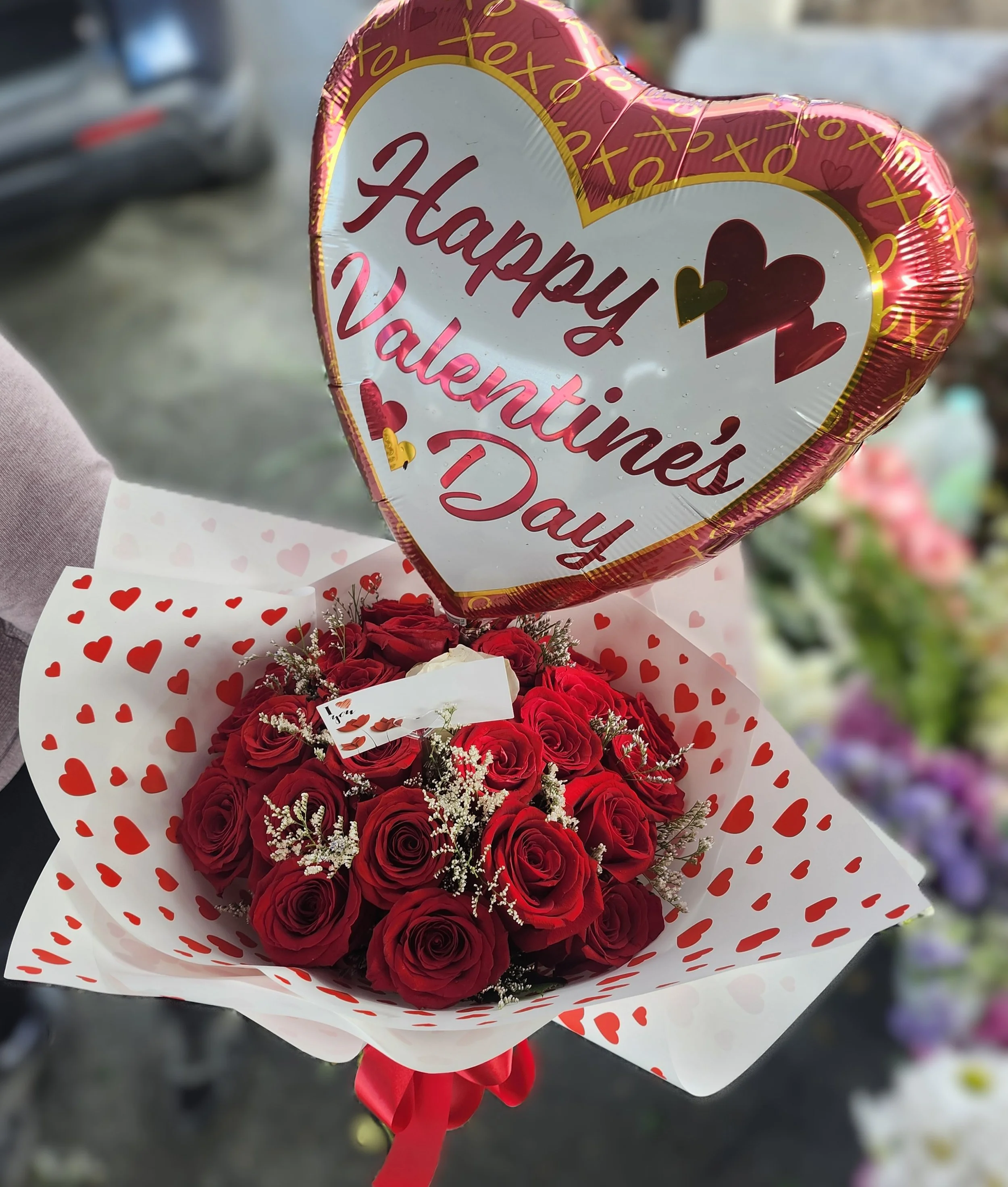 A bouquet of red roses with white baby's breath, wrapped in white paper with red hearts, and a heart-shaped balloon with a gold border that says 'Happy Valentine's Day' in red and pink letters.