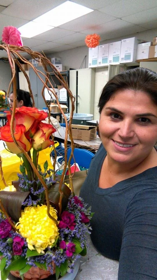 Woman smiling in a room with office supplies, taking a selfie with a flower arrangement featuring roses and other colorful flowers.