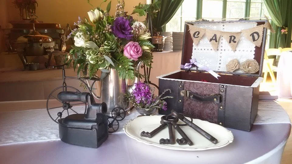A table decorated with a bouquet of purple and white flowers, vintage keys on a white tray, a black vintage-style iron, and an open decorative chest with a banner spelling "E A R D." in a room with green curtains and sunlight.