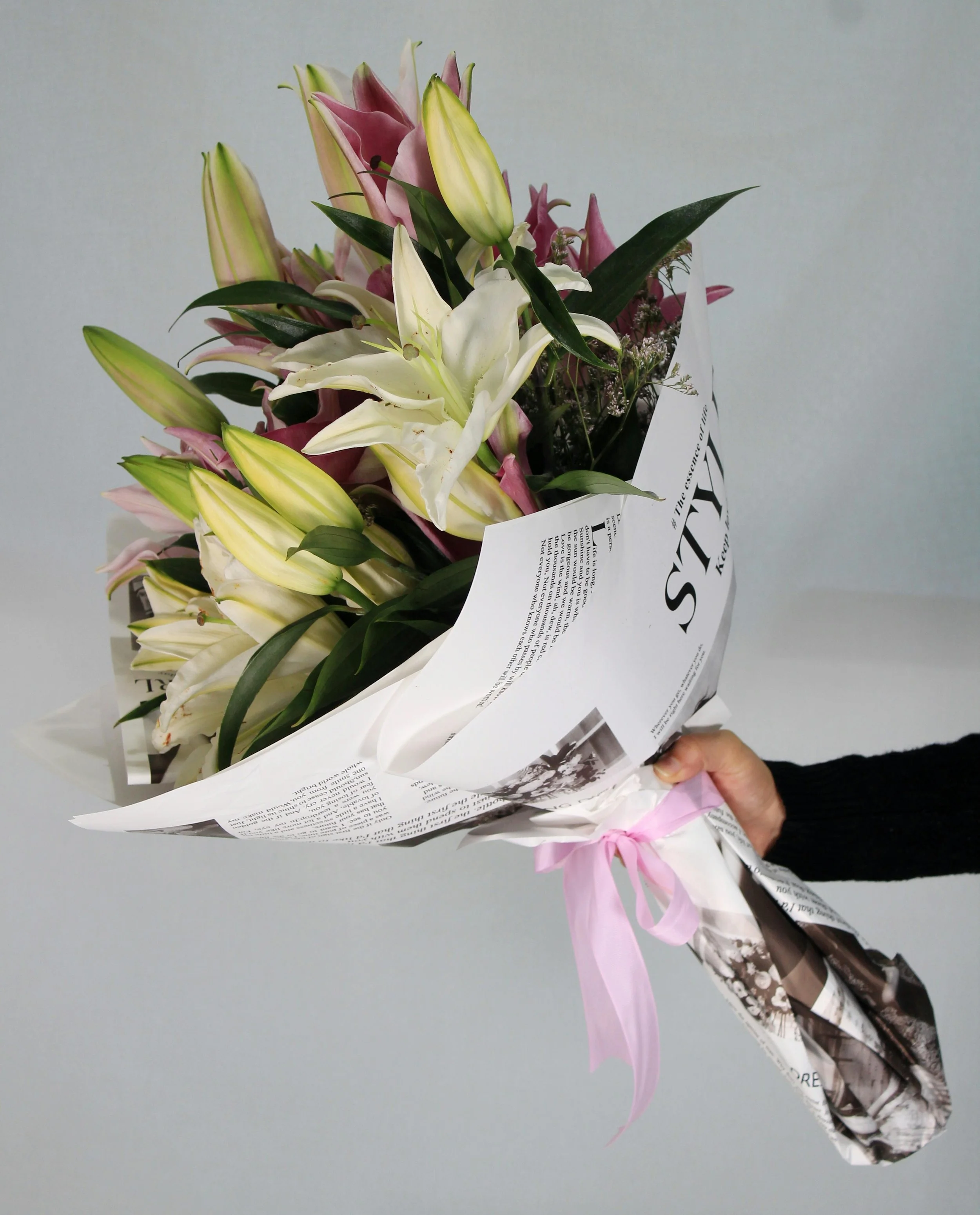 A person holding a bouquet of pink and white lilies wrapped in newspaper with a pink ribbon.
