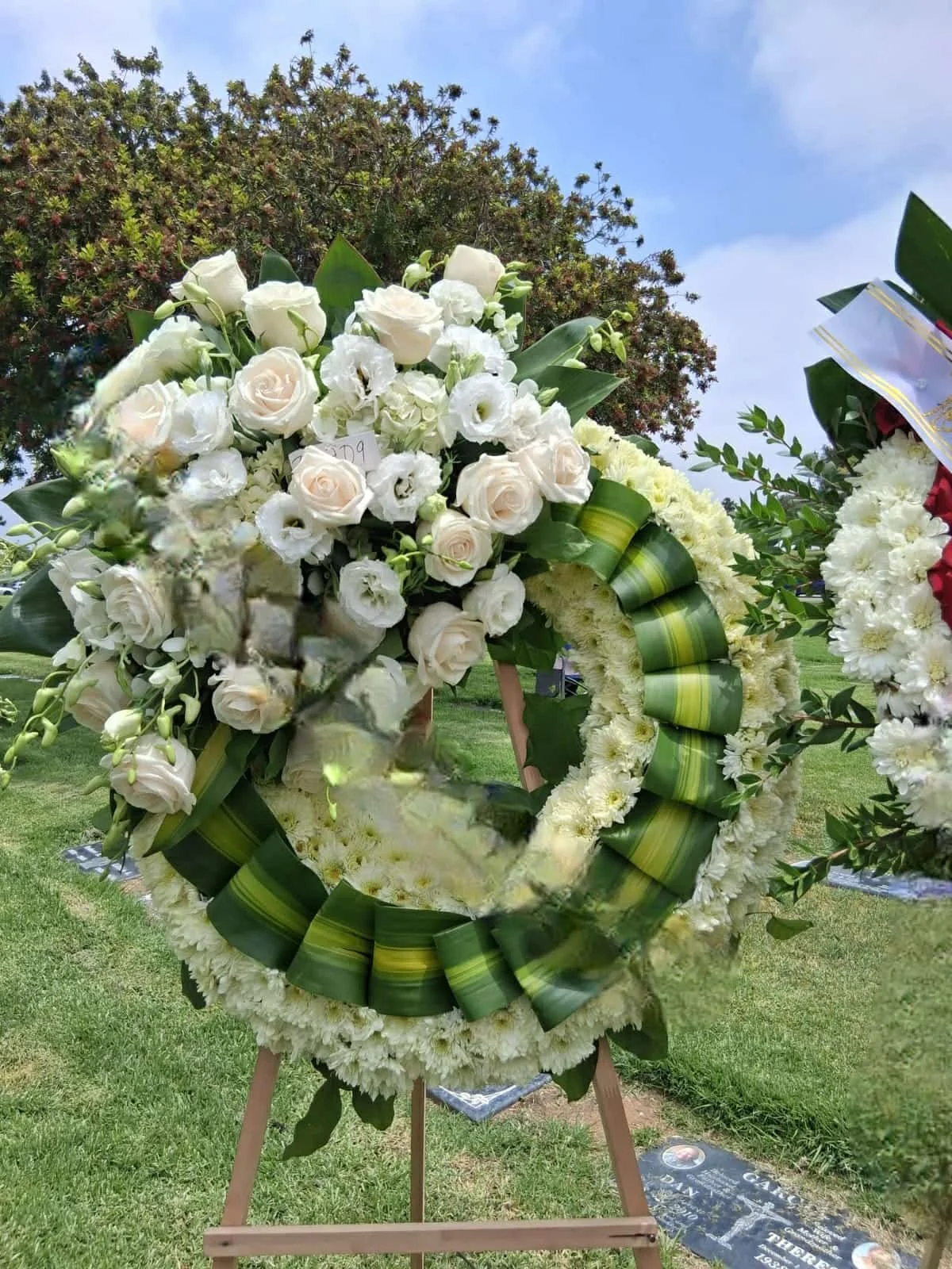 A funeral flower wreath made of white roses, white lilies, and white carnations on a wooden stand outdoors, with a grassy area and cloudy sky in the background.