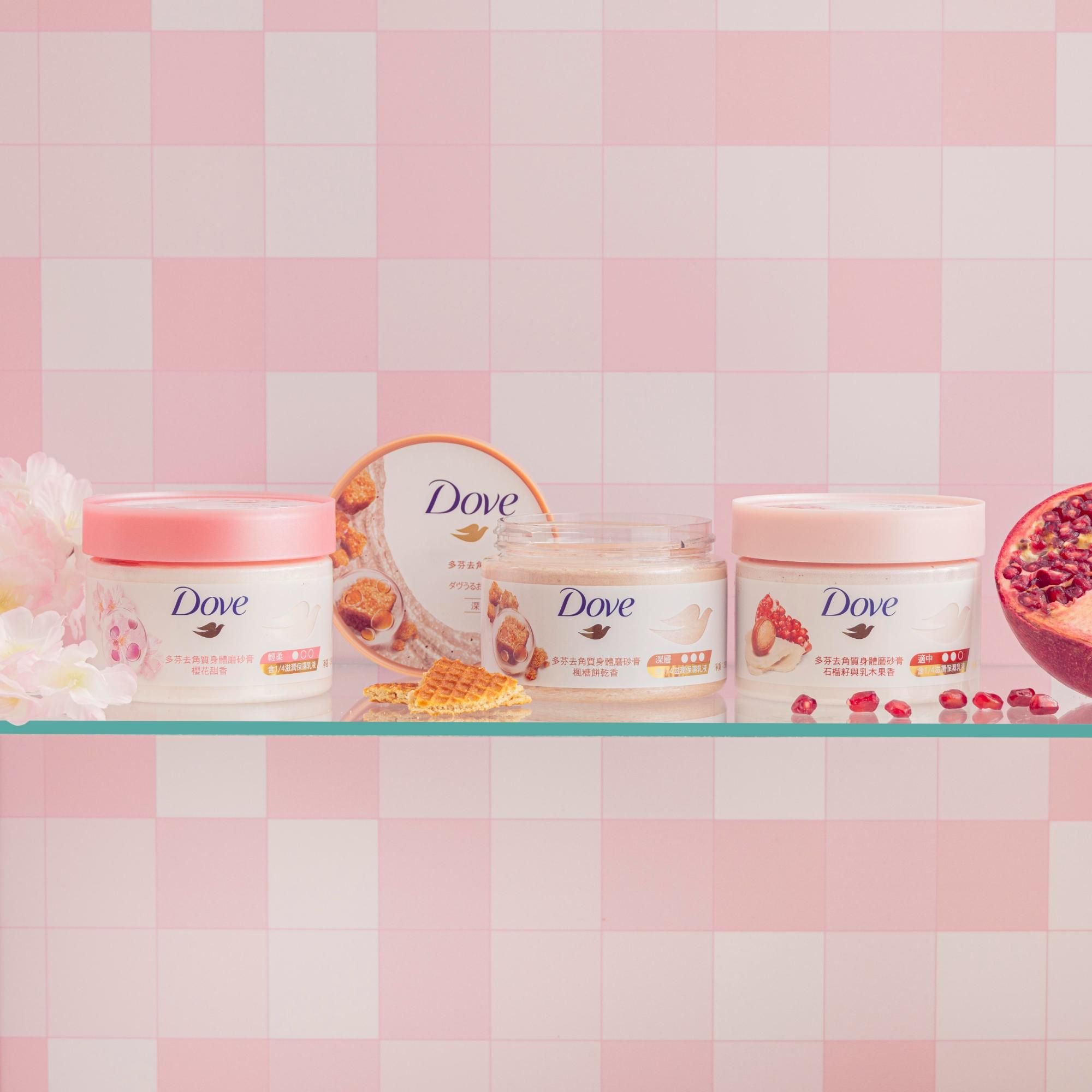 Various Dove skincare products with pink lids, including body scrub and body cream, displayed on a glass shelf against a pink checkered background, with flowers and pomegranate on either side.