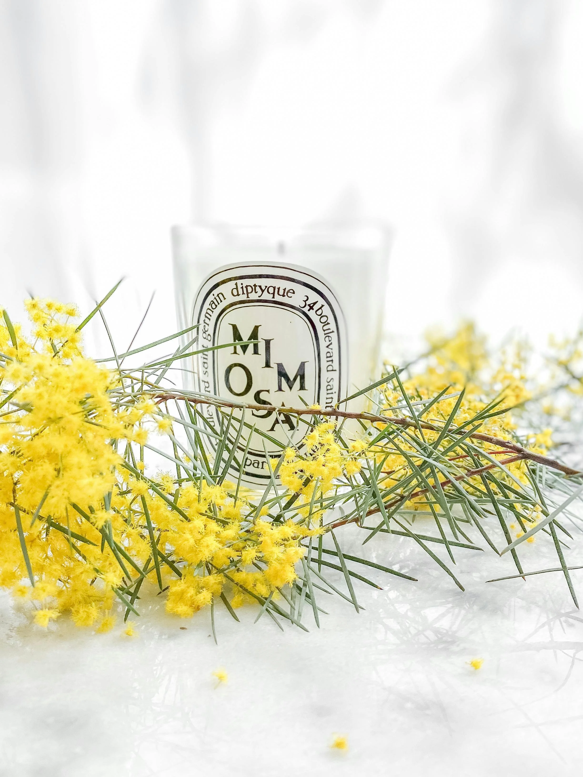 A white candle with a black and white label, surrounded by yellow flowers and green foliage on a white surface.