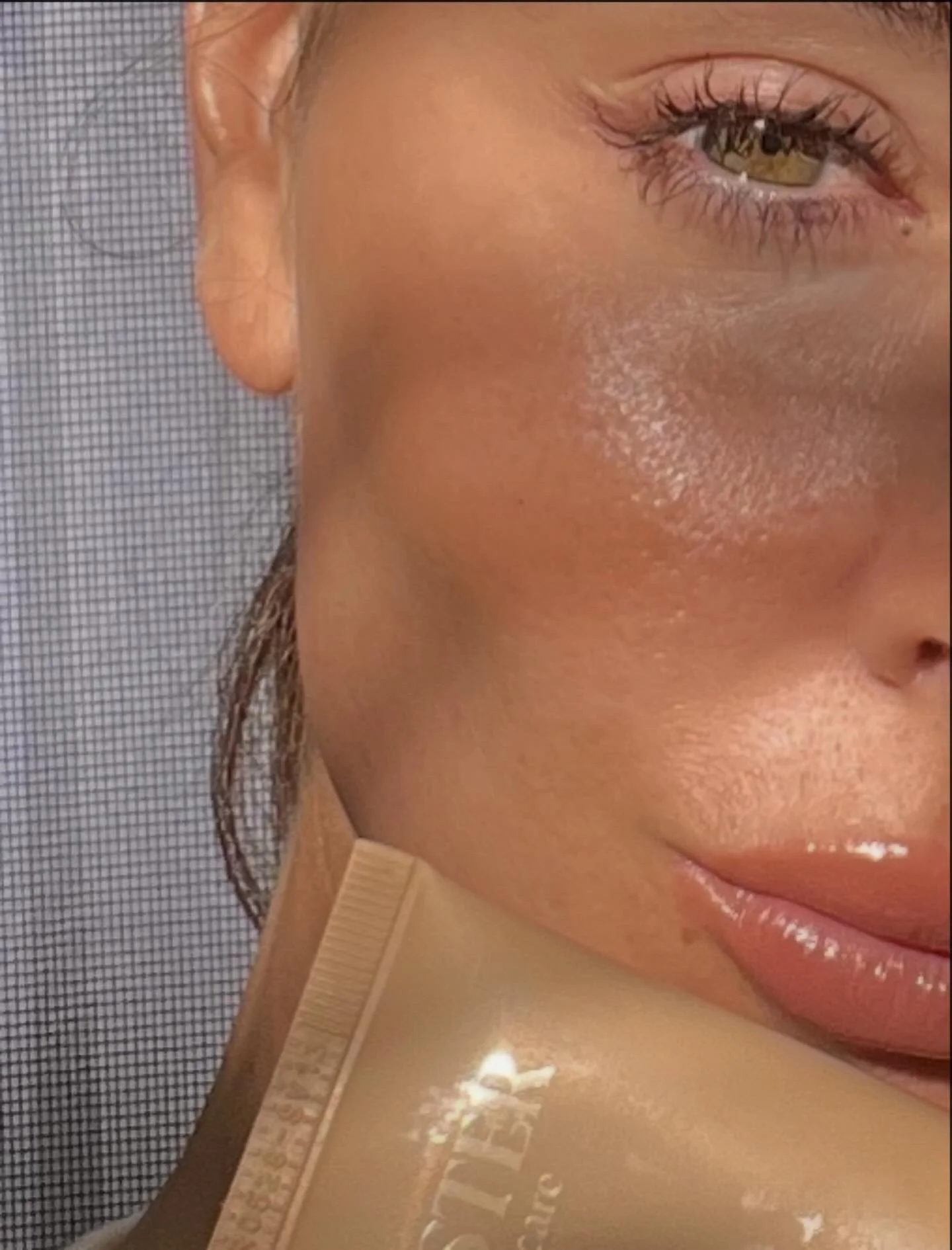 A close-up partial view of a woman's face showing her eye, cheek, nose, and lips, with a beige-colored skincare or cosmetic product near her chin.