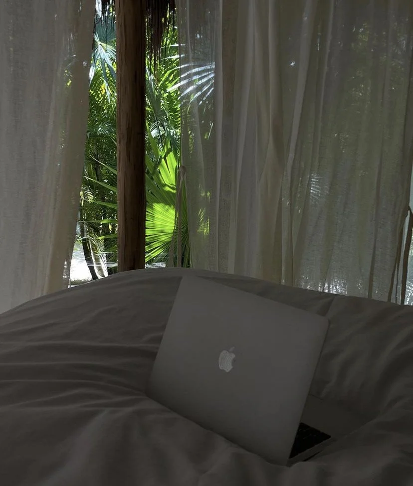 A MacBook placed on a soft bed or couch with beige or light-colored bedding, in front of a window with sheer white curtains. Outside the window, there are lush green tropical plants and palm leaves suggesting a tropical or jungle setting.