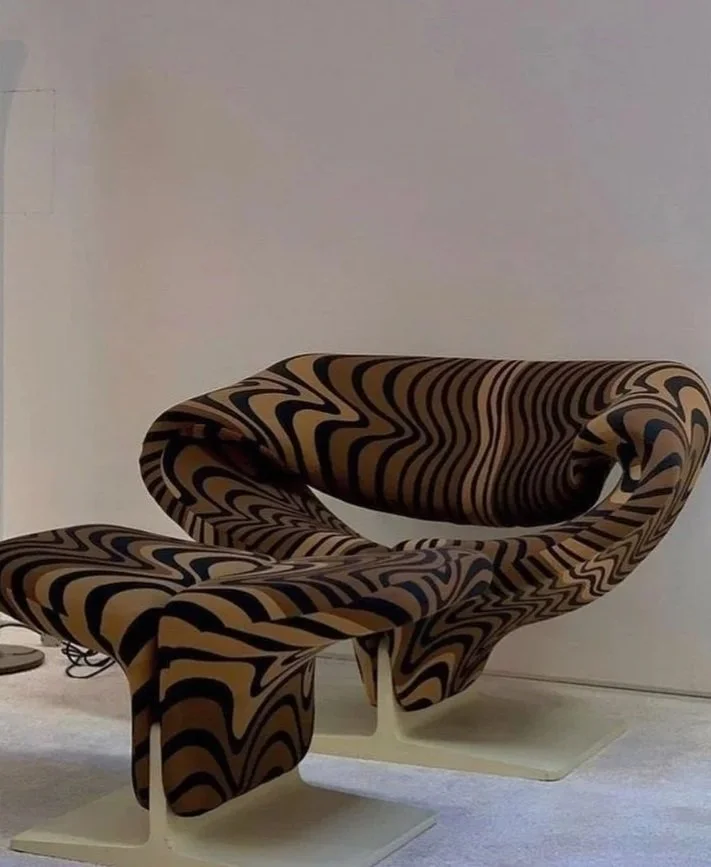 A modern chair with a matching footrest, both covered in a black and beige abstract patterned fabric, placed against a plain wall.