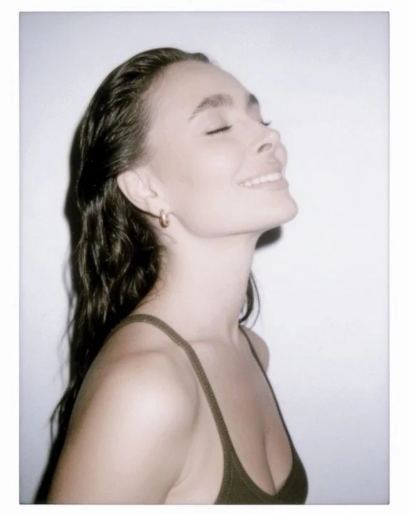 A young woman with long, wavy brown hair smiling with her eyes closed, facing slightly to the left, against a plain white background.