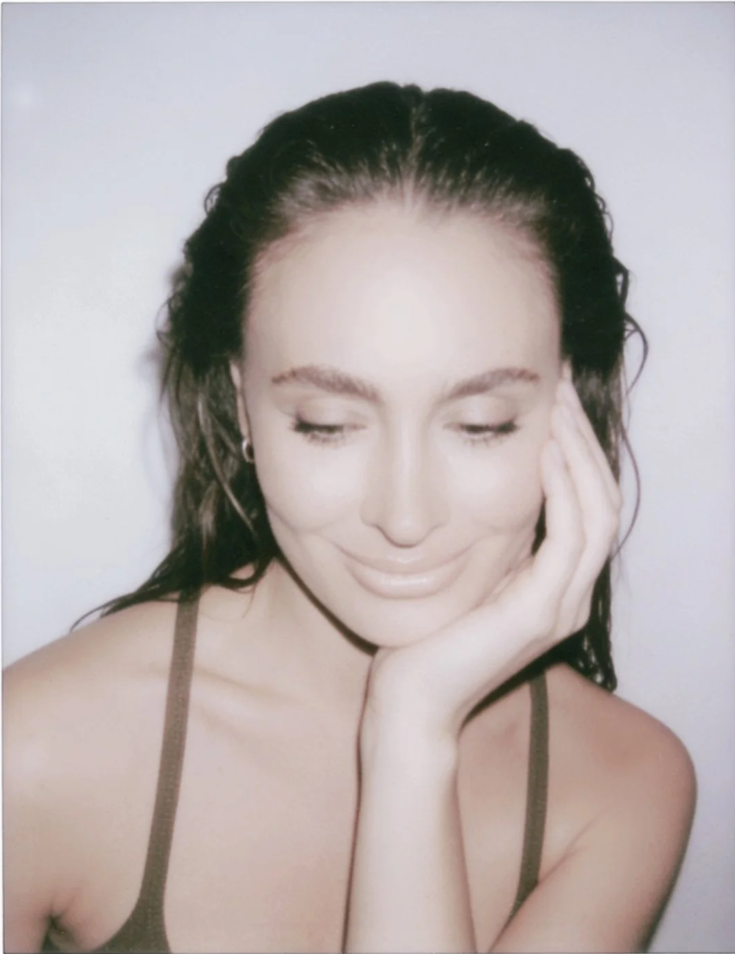 A woman with dark wet hair, closed eyes, and a gentle smile, resting her face on her hand against a plain background.