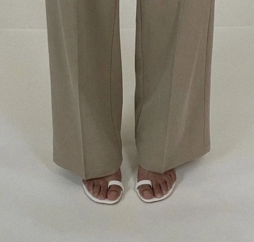 Close-up of a person wearing beige wide-leg pants and white open-toe sandals, standing on a white surface.
