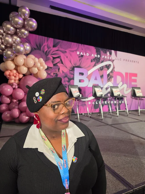 Brittney Sherell wearing a black beret and glasses in front of a pink Baldie Con backdrop