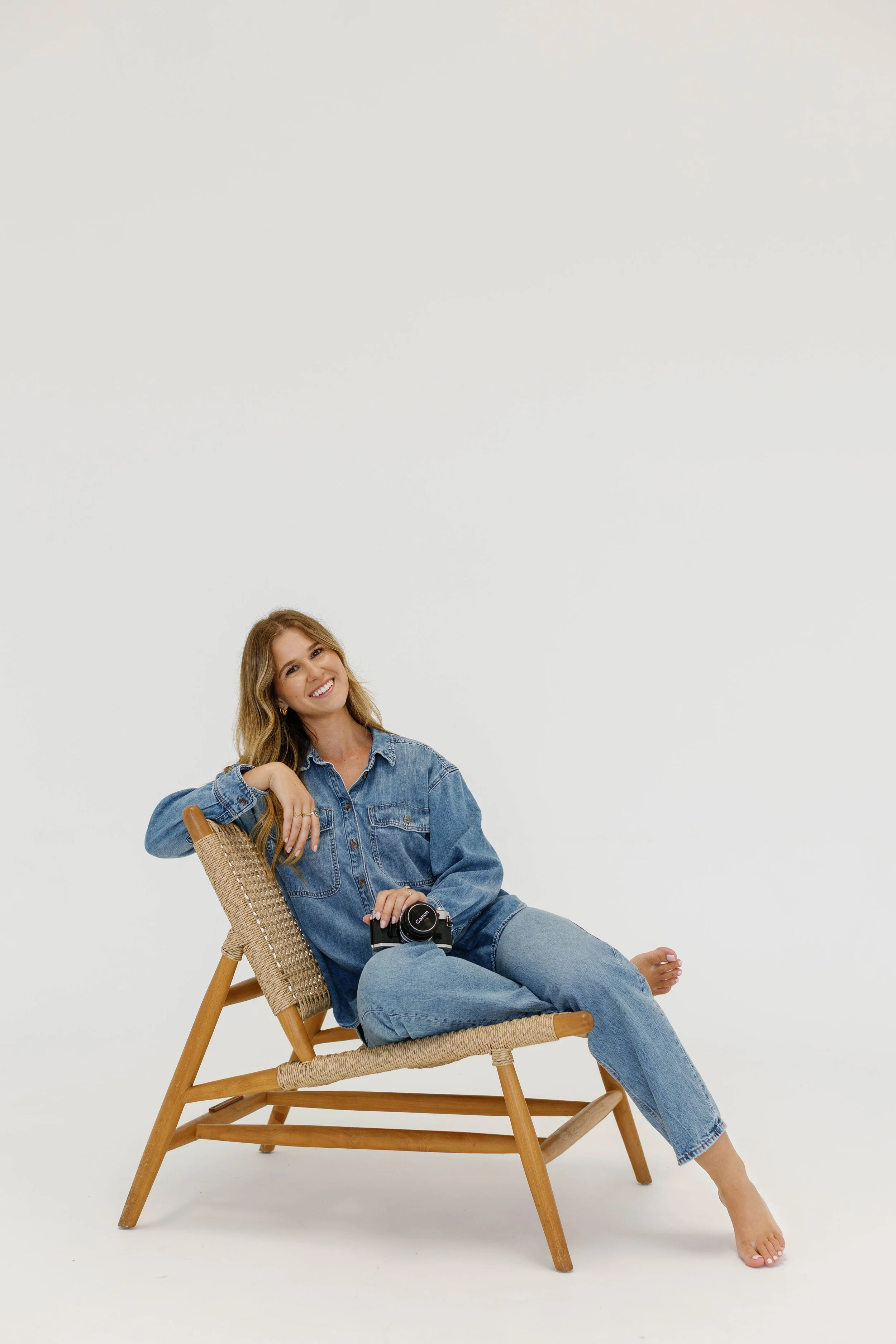 A woman with long blonde hair sitting on a wooden chair with woven seat and back, smiling, dressed in a denim jacket and jeans, holding a vintage camera, against a plain white background.