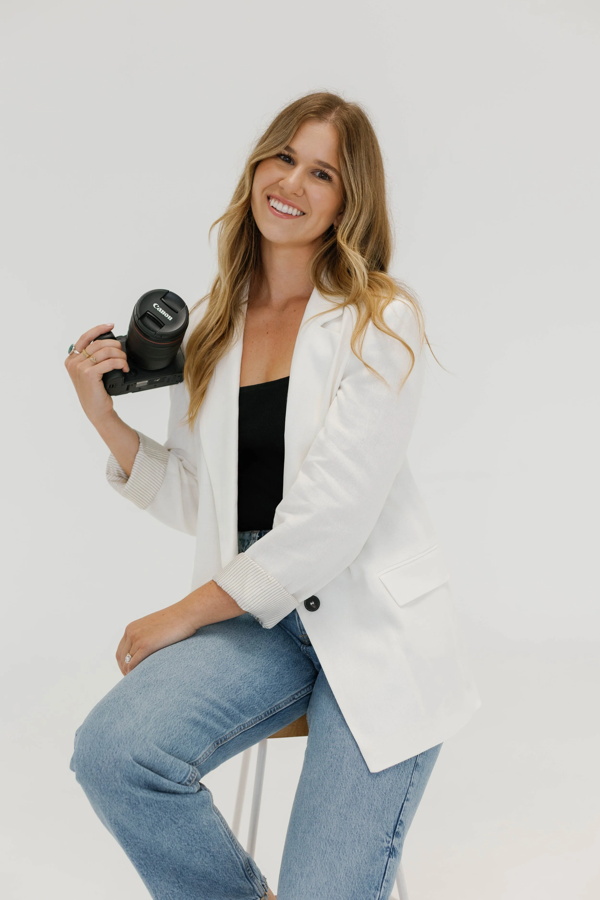 Smiling woman with long, wavy blonde hair sitting on stool, holding DSLR camera, wearing white blazer over black top and blue jeans, against plain white background.
