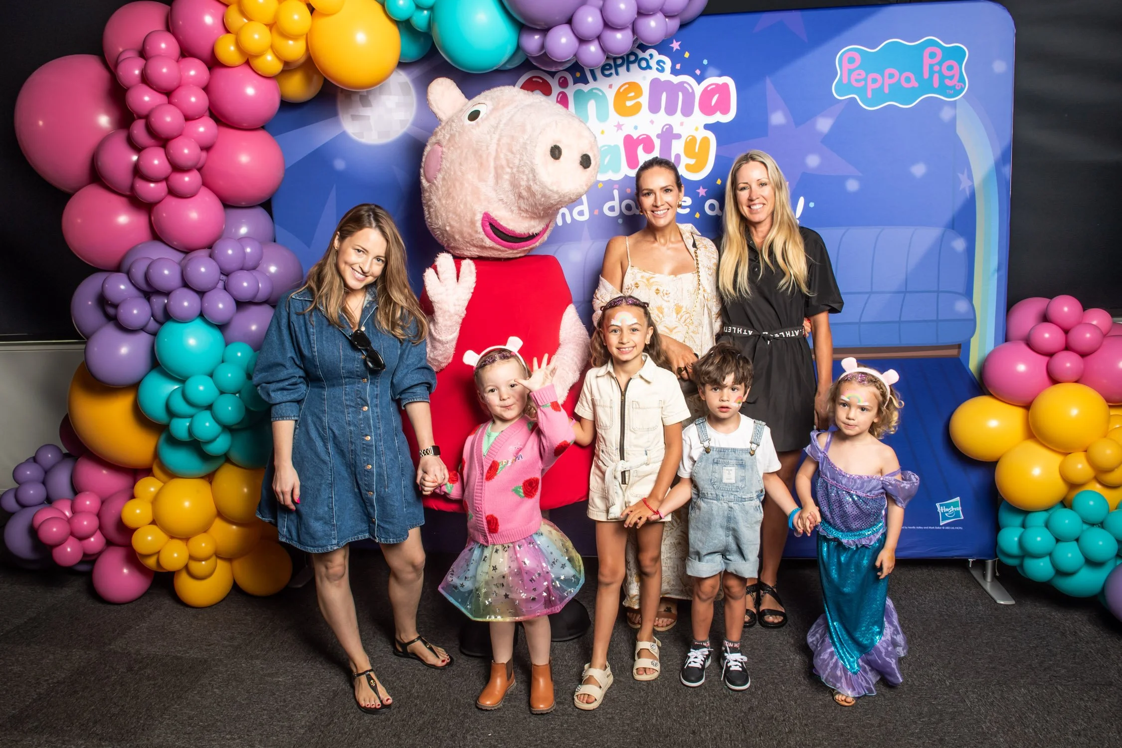 Family posing with Peppa Pig mascot at a Peppa Pig event, with children and adults smiling, pink and purple balloons, and a Peppa Pig backdrop.