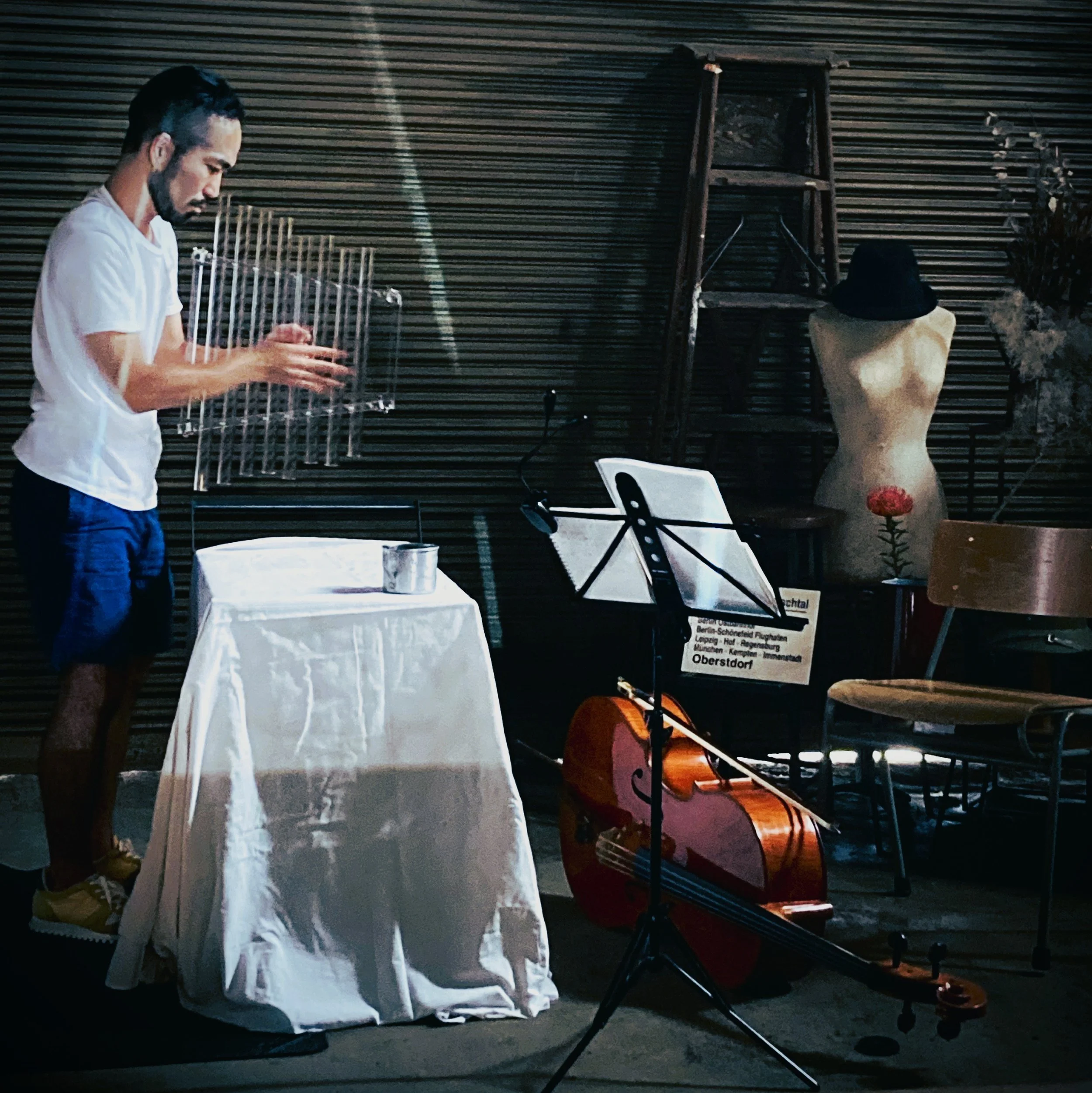 ステージにいる男性が何らかの金属のオブジェを持っている。背景には木製の棚とマネキン、椅子、楽器や譜面台が置かれている。