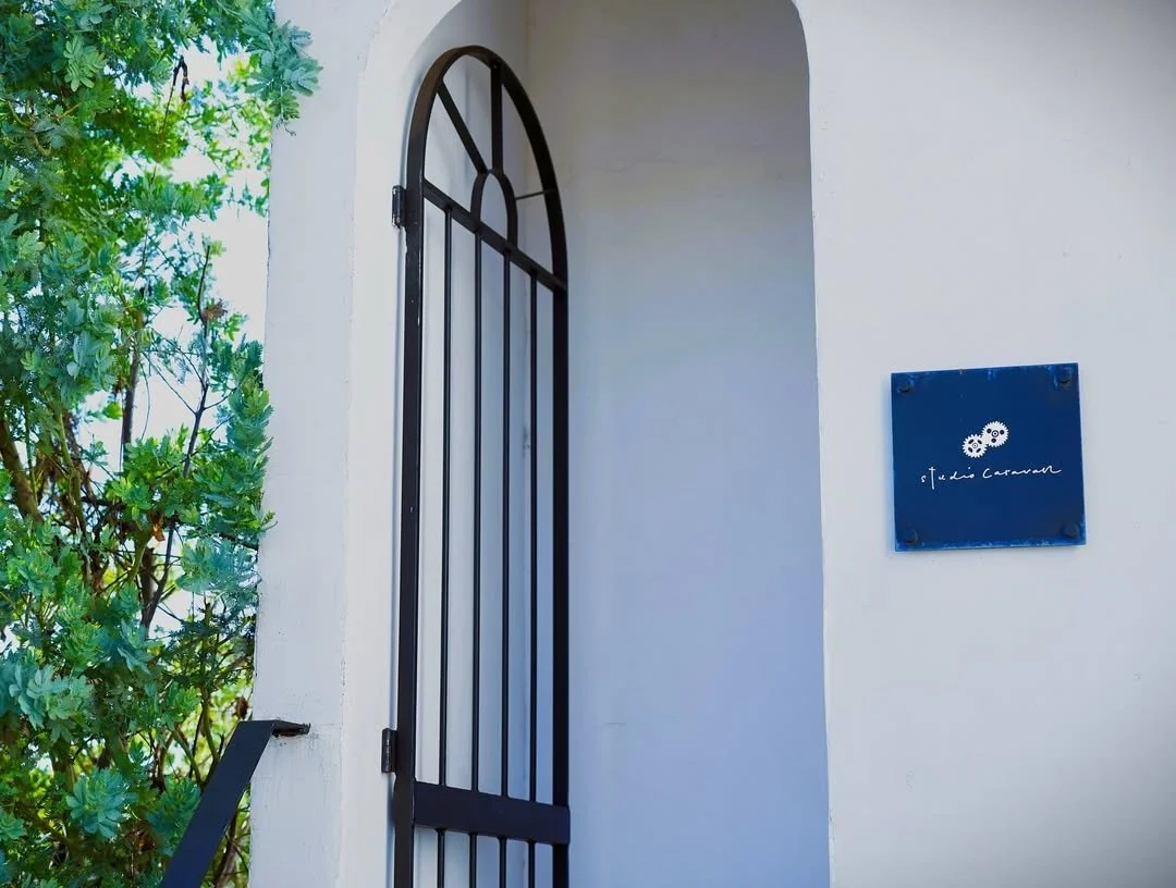 白い壁に黒いアーチ型の鉄製門と青い看板のstudio caravanの入口