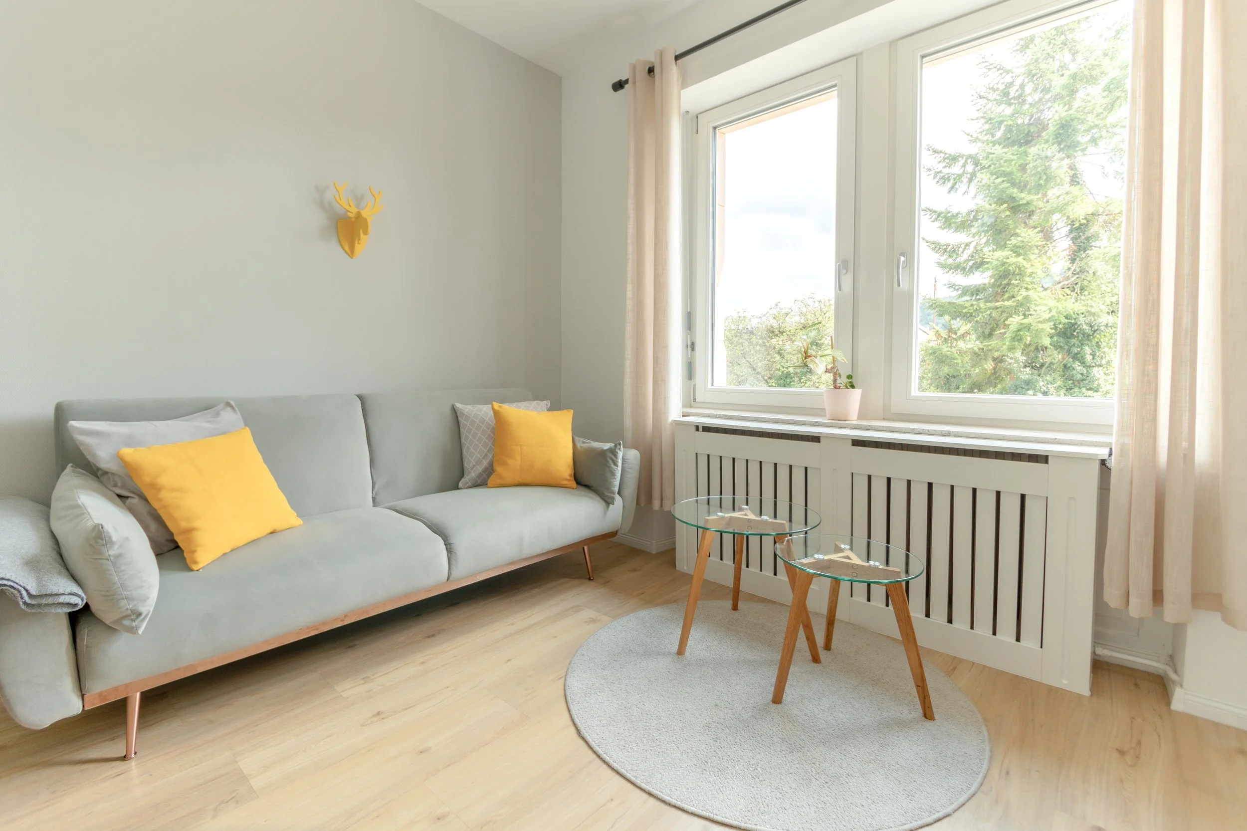 Helles Wohnzimmer mit gemütlichem Sofa, freundlichen Gelb-Akzenten und großem Fenster mit Blick ins Grüne – ideal zum Entspannen.
