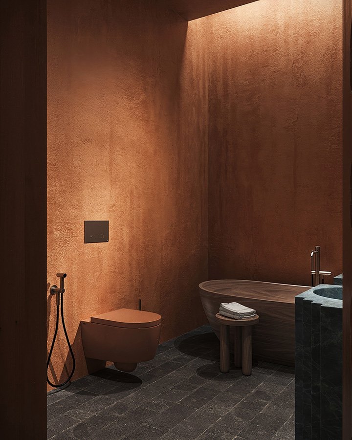A modern bathroom with copper-colored textured walls, a hidden toilet with a wall-mounted flush panel, a freestanding wooden bathtub with towels, a small wooden stool, and a dark stone sink with a faucet.