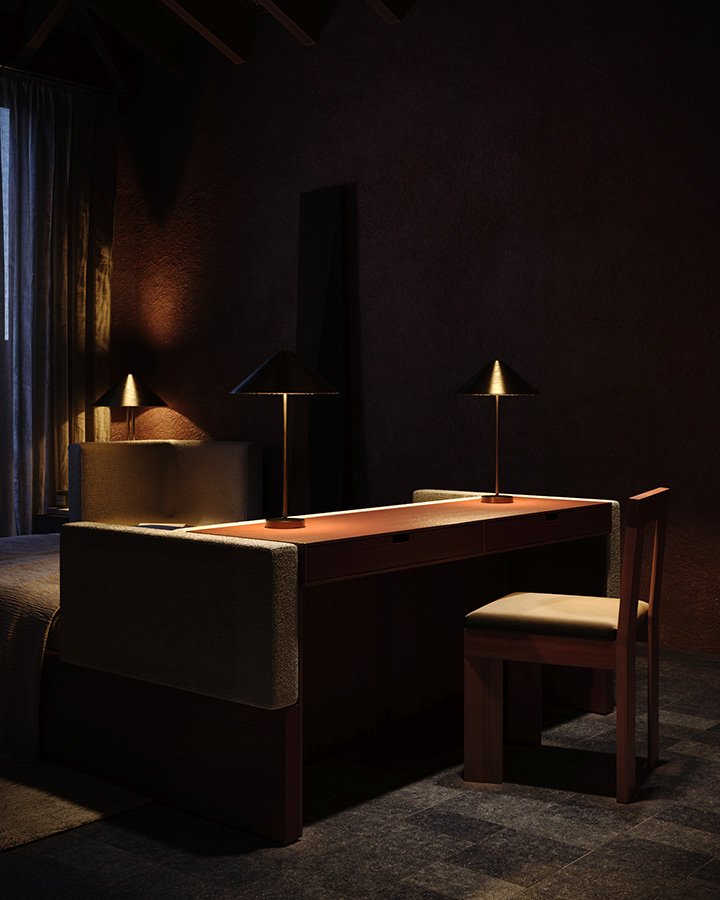 Dimly lit room with a wooden desk, two desk lamps, a cushioned chair, and a bed partially visible on the left, with dark walls and curtains.