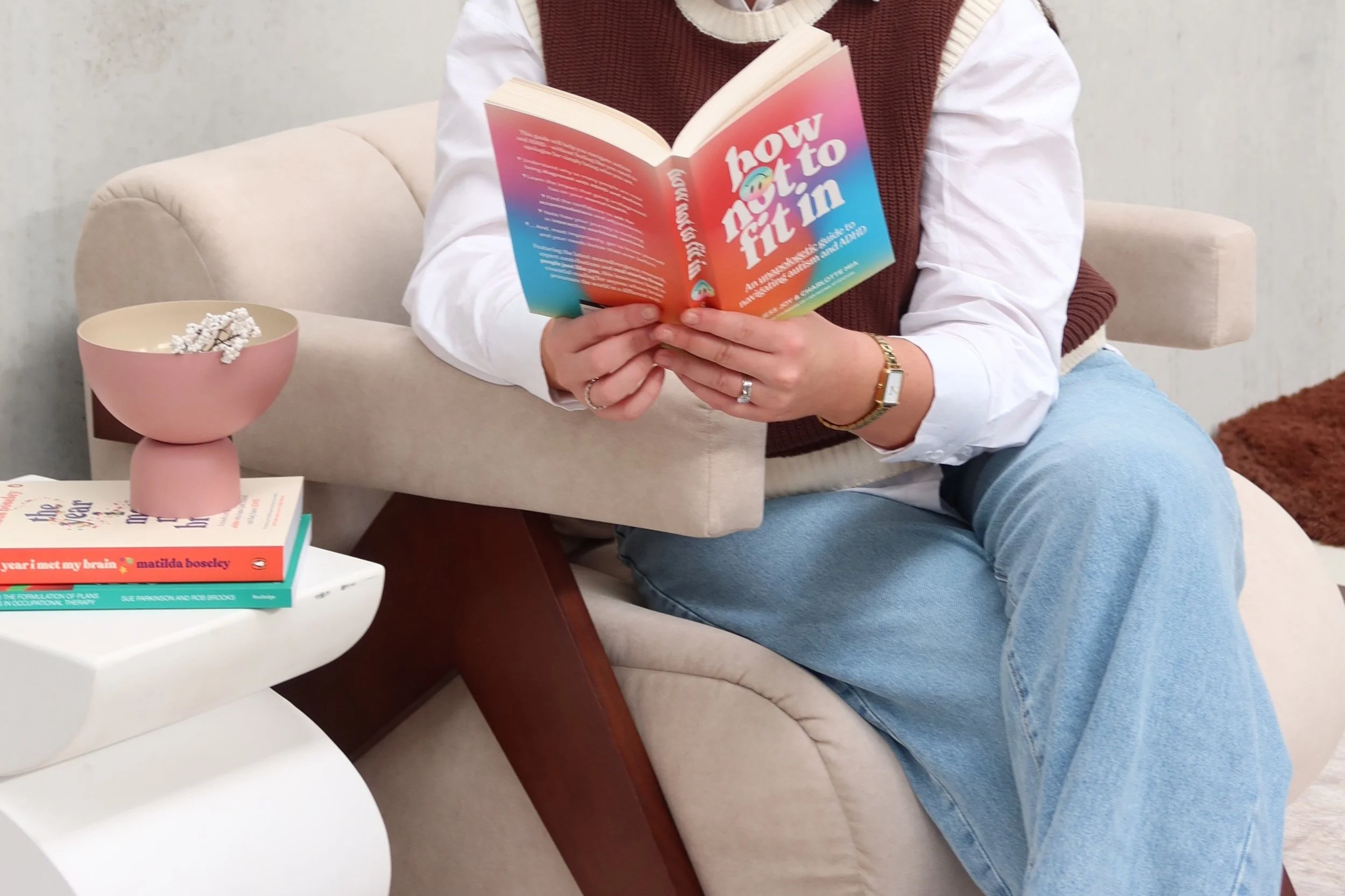 Person sitting on a beige sofa reading a colorful book titled 'How Not to Fit In'. They are wearing a white shirt, a brown vest, and light blue jeans. Next to them is a white side table with two books on it, a pink bowl with popcorn, and a gold wristwatch.