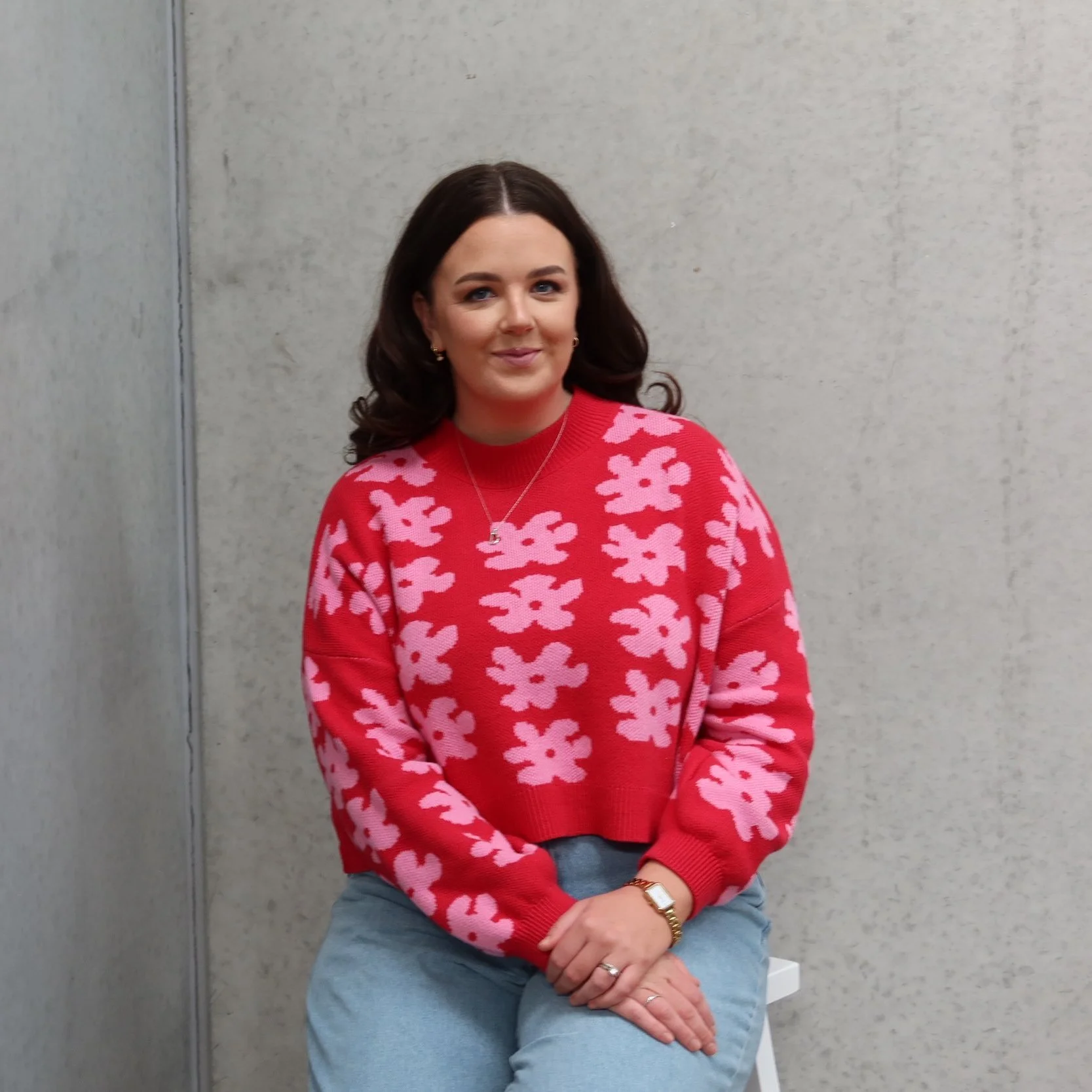 A woman with long wavy brown hair seated against a plain concrete wall, wearing a red sweater with pink floral patterns, light blue jeans, and accessorized with a watch, a ring, earrings, and a delicate necklace.