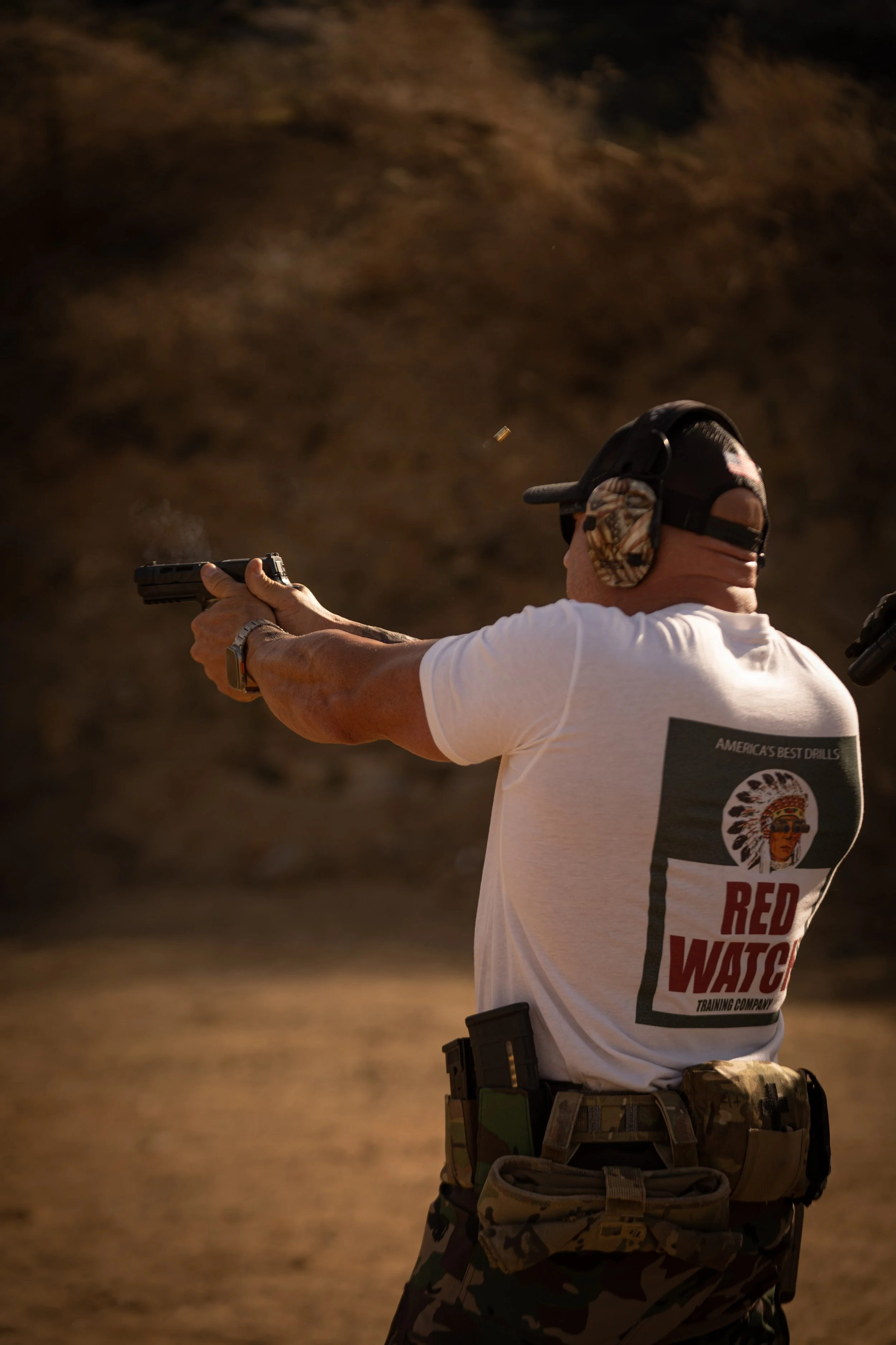 A man aiming a handgun at an outdoor shooting range, wearing ear protection and camouflage pants, with a white t-shirt that reads "America's Best Drills" and "Red Watch Training Company."