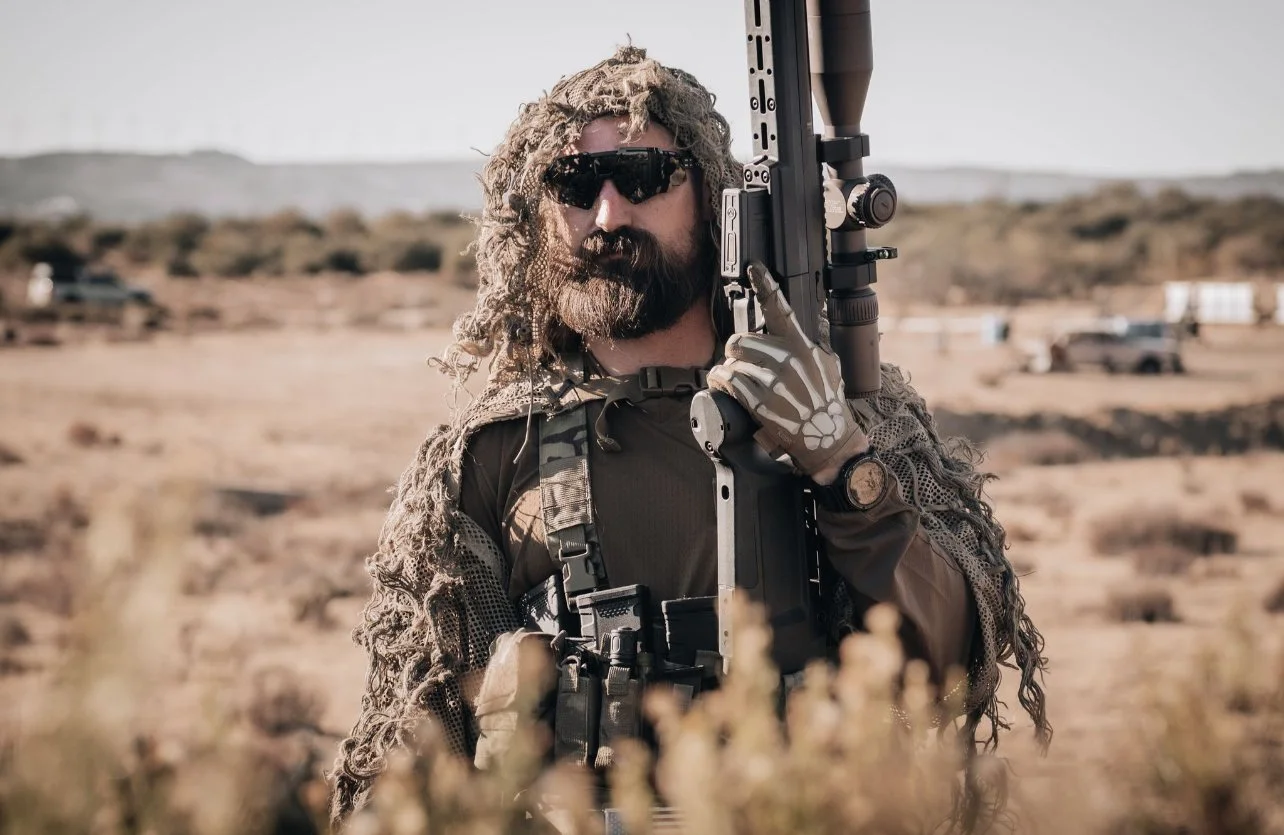 A man with a beard wearing sunglasses, military gear, and camouflage clothing, holding a sniper rifle in a desert landscape.