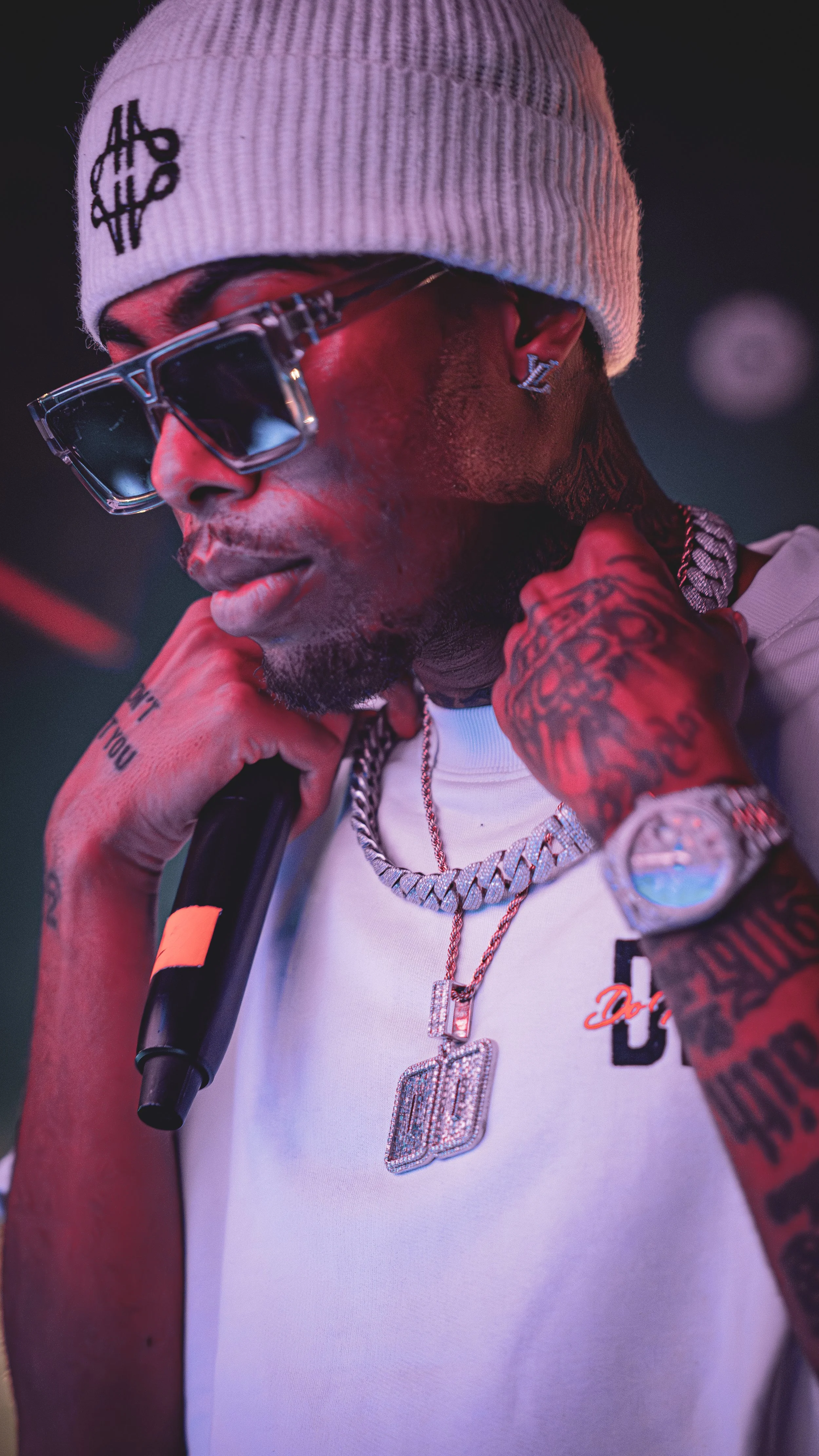 A man wearing a white beanie, large sunglasses, a white T-shirt, and heavily tattooed arms. He has jewelry including a chain necklace with pendants, a large watch, and earrings. The man holds a microphone near his chin and appears to be in a performa