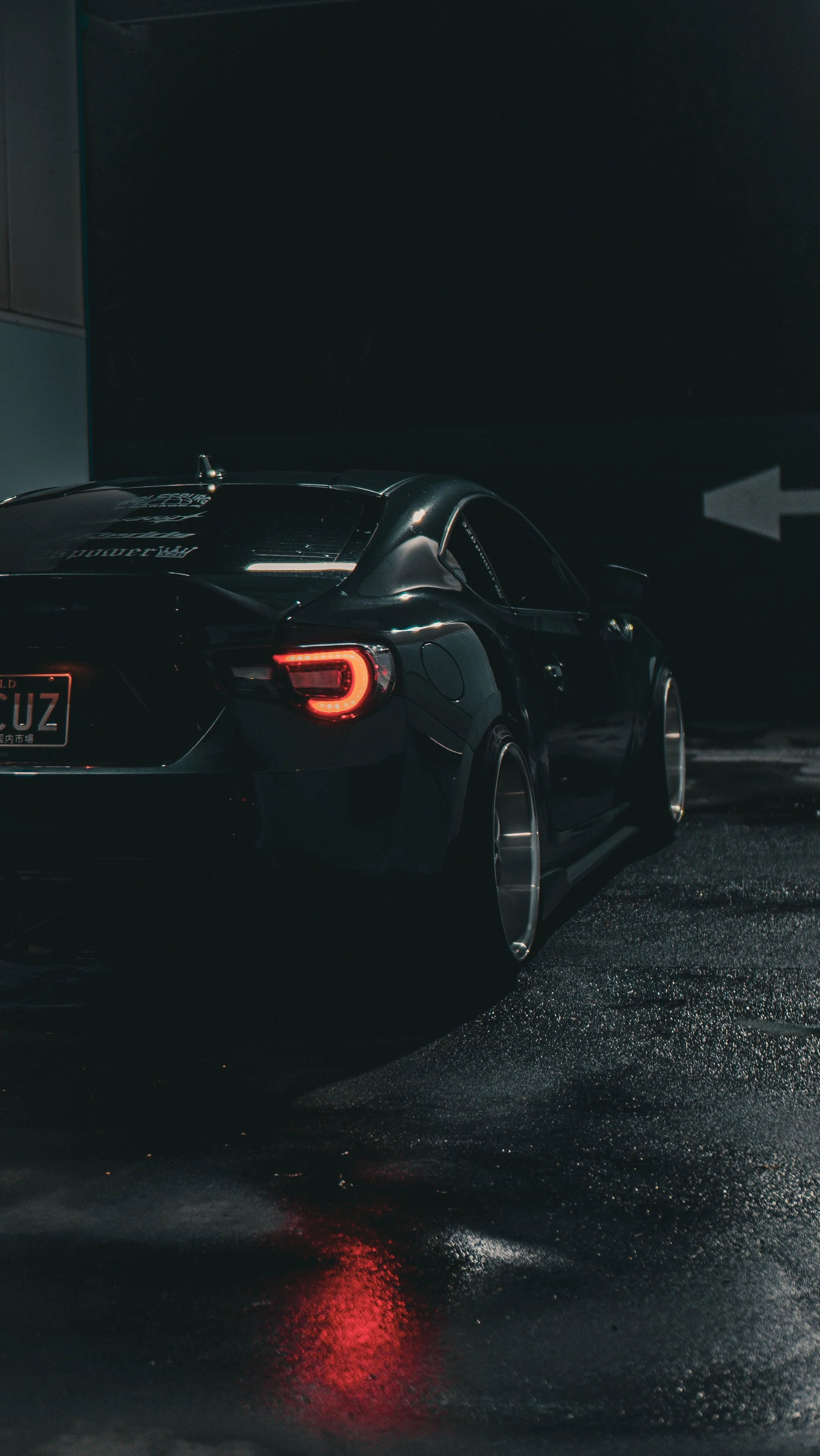 A sleek black sports car with bright tail lights parked in a dark, wet parking garage with reflections on the ground.