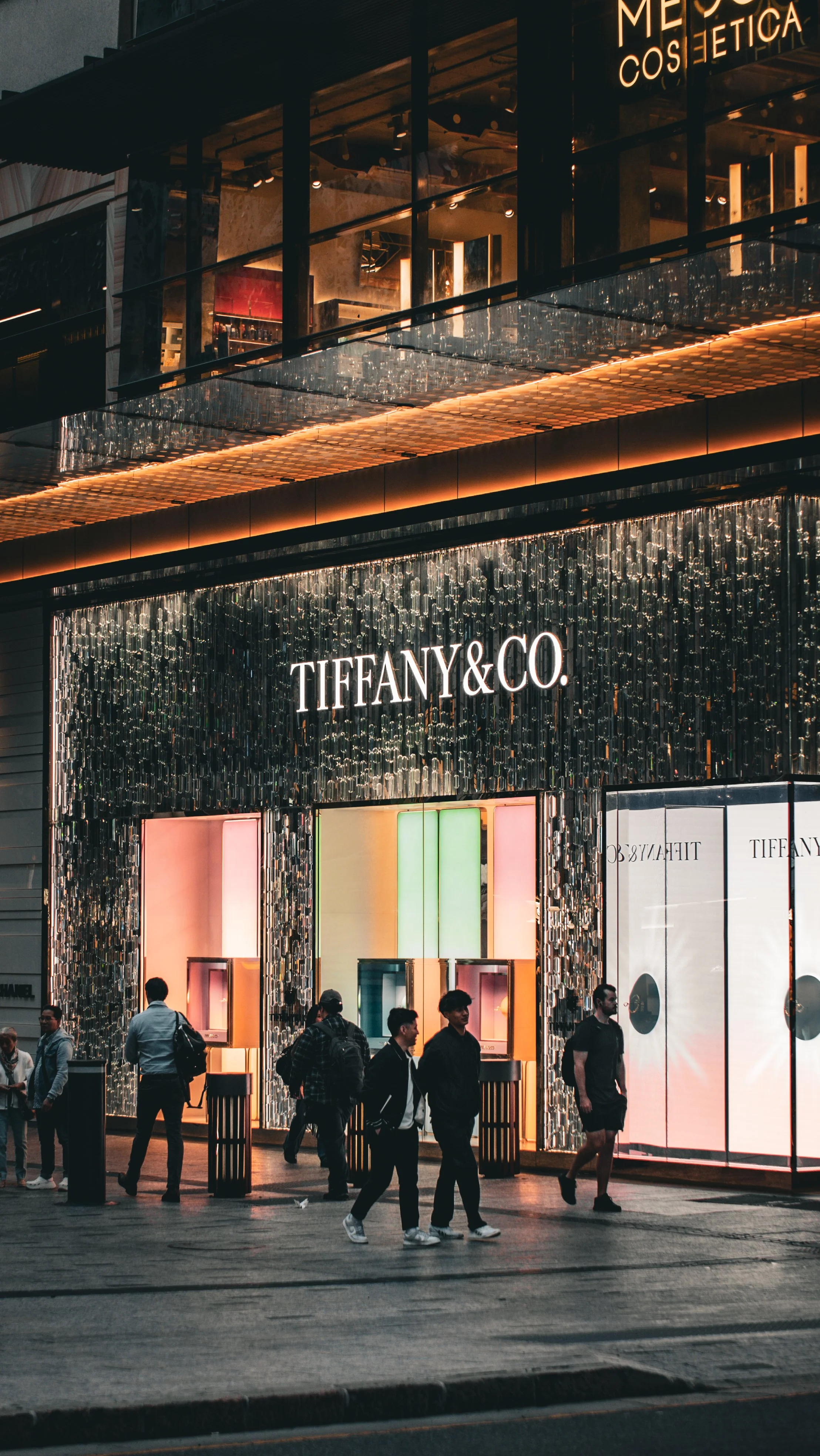 People walking past the Tiffany & Co. storefront illuminated at night, with a shiny, reflective exterior and colorful display lights