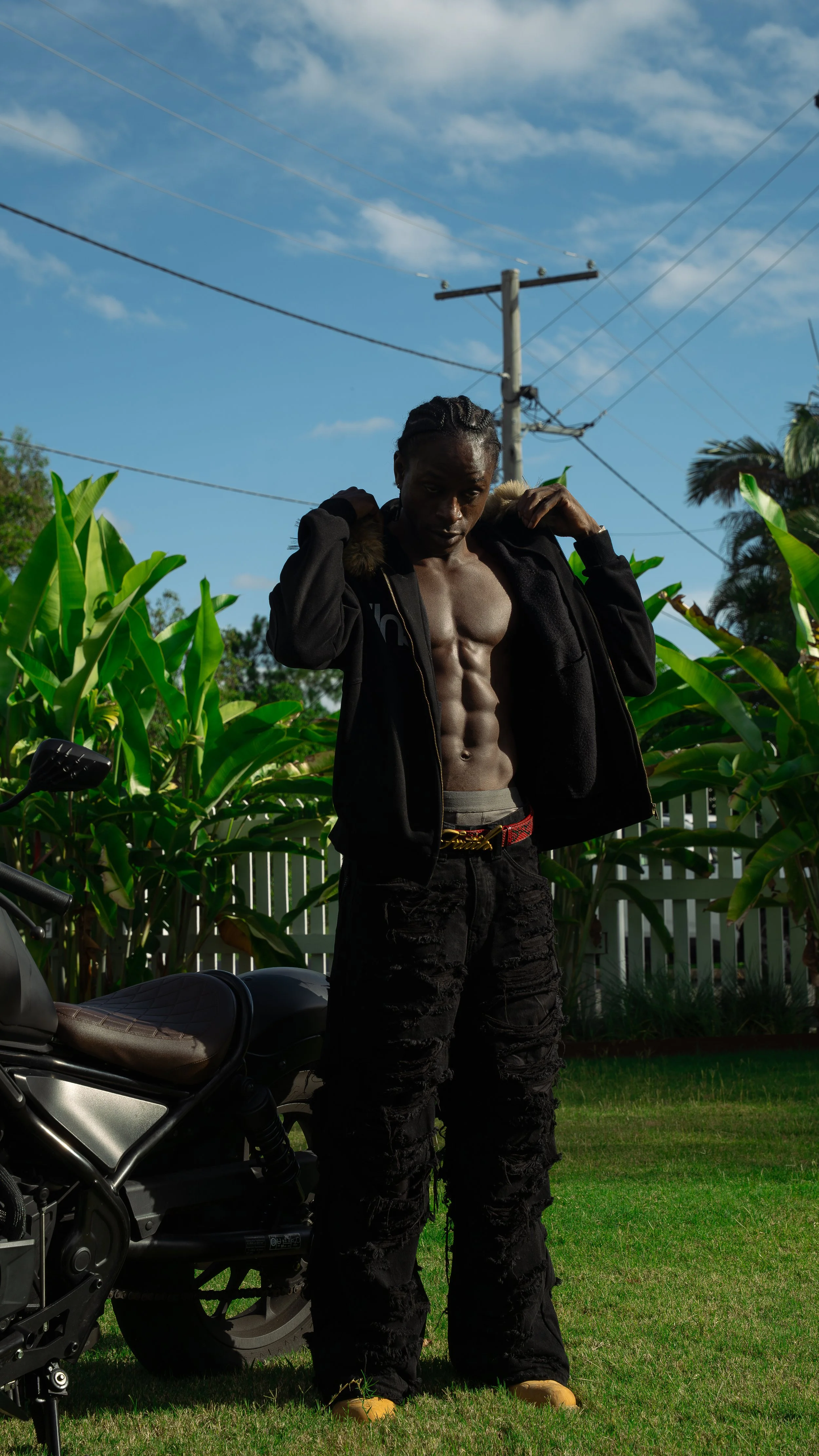 A shirtless man with a muscular abdomen standing outdoors next to a motorcycle. He's wearing black ripped pants and a black jacket with a fur-lined hood, posing with his hands on his shoulders. There are green plants and a fence in the background und