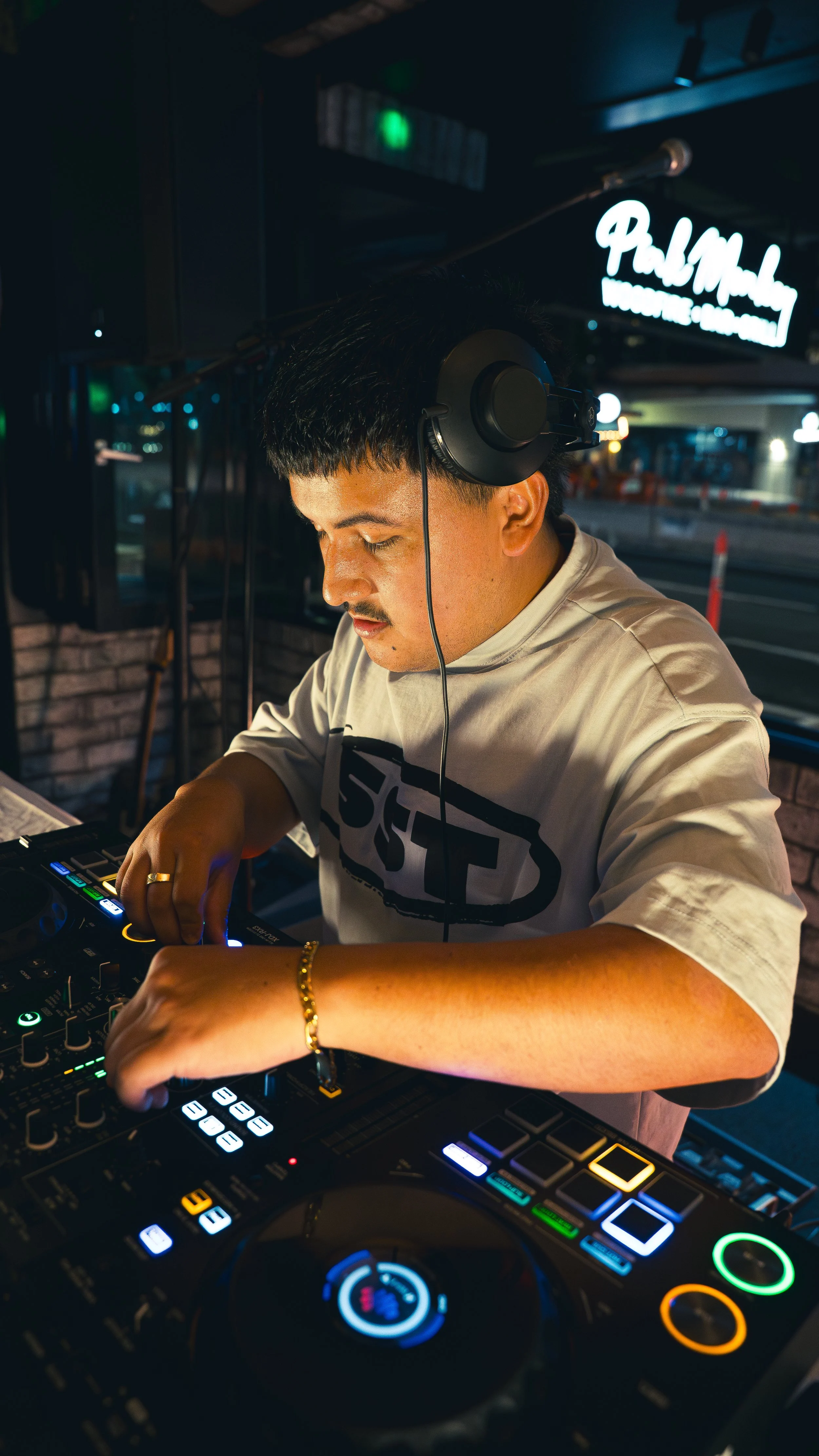 A male DJ wearing headphones mixes music at a club or bar, with neon signage and a city street visible in the background.