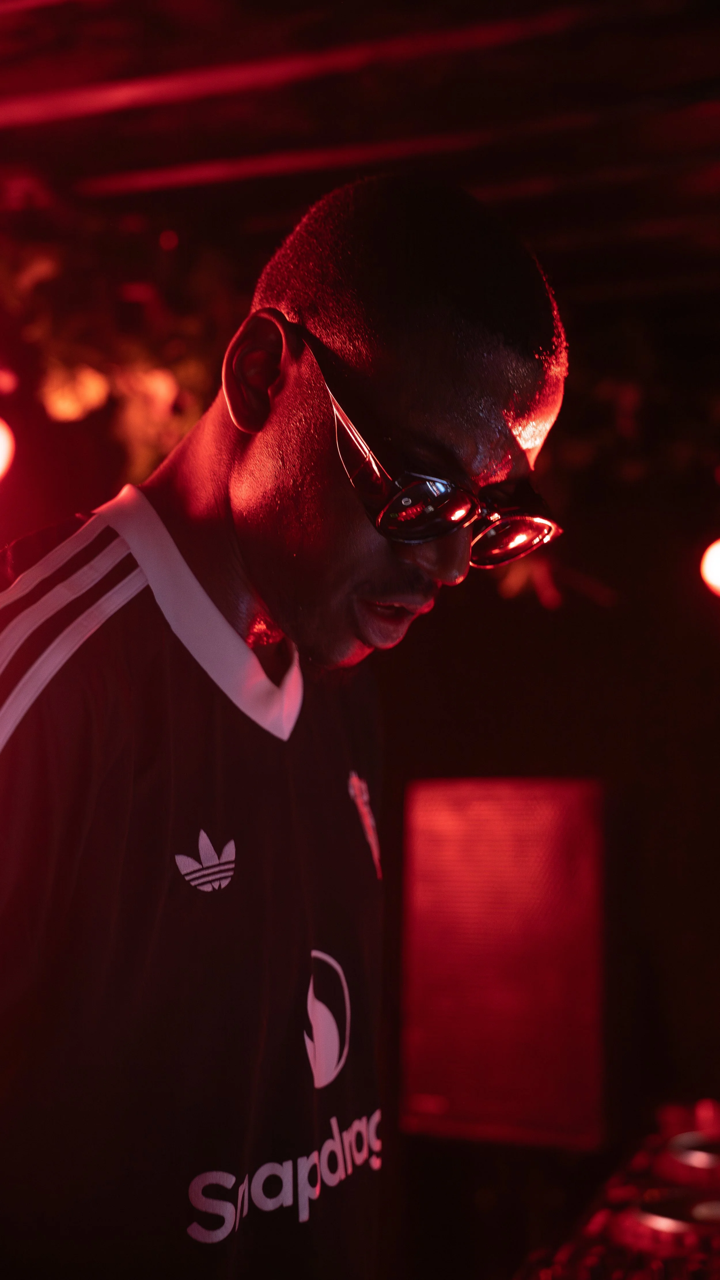 A man wearing sunglasses and a black Adidas jacket with white stripes, illuminated by red lighting, appears to be DJing at a club or event.
