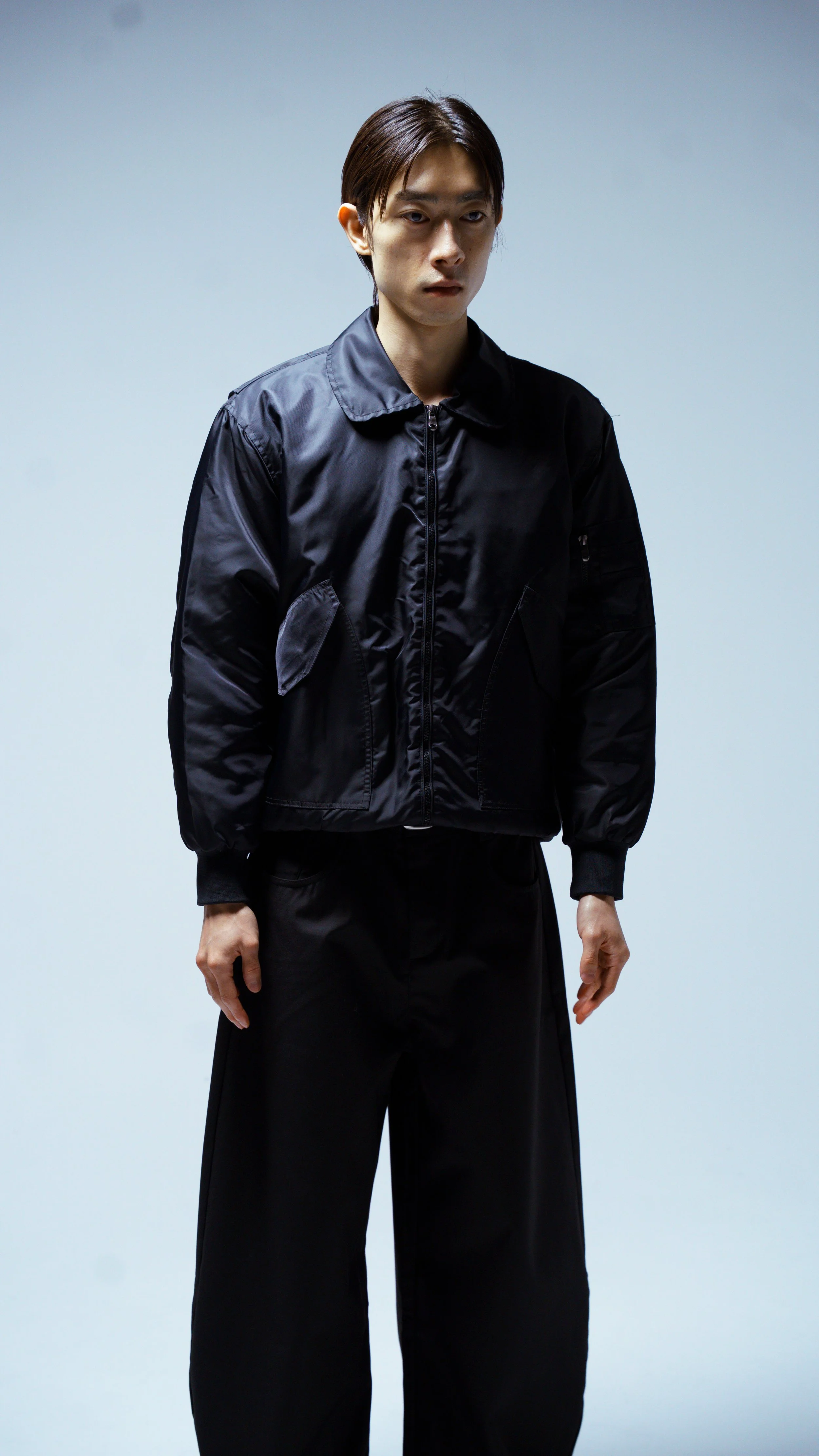 A person wearing a black bomber jacket and black wide-leg pants standing against a plain light background.