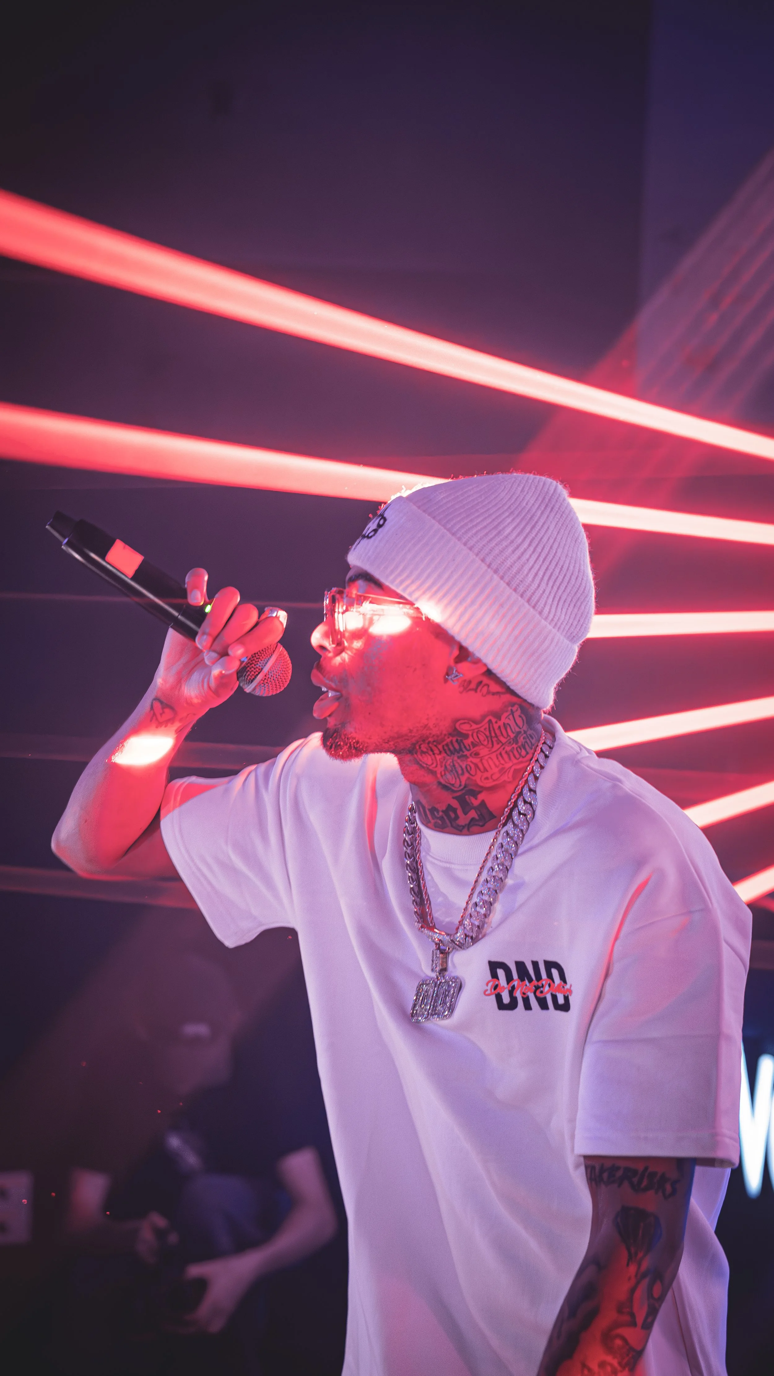A man performing on stage, wearing a white beanie, glasses, and a white t-shirt. He has tattoos on his neck and arms, and is holding a microphone while singing or rapping. red laser lights are in the background.