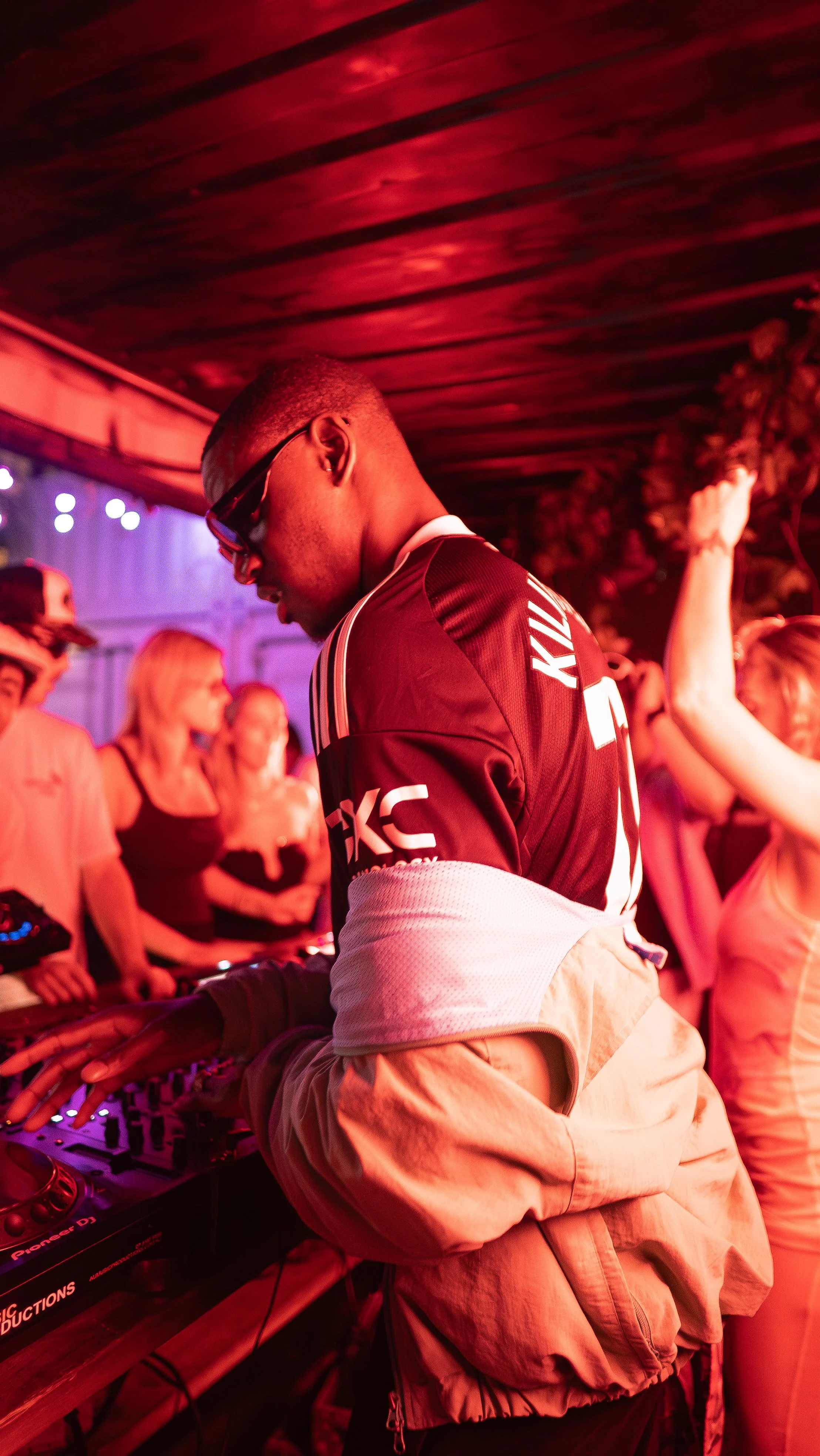A DJ in a black and white sports jacket and sunglasses mixes music at a party with a crowd dancing around him.