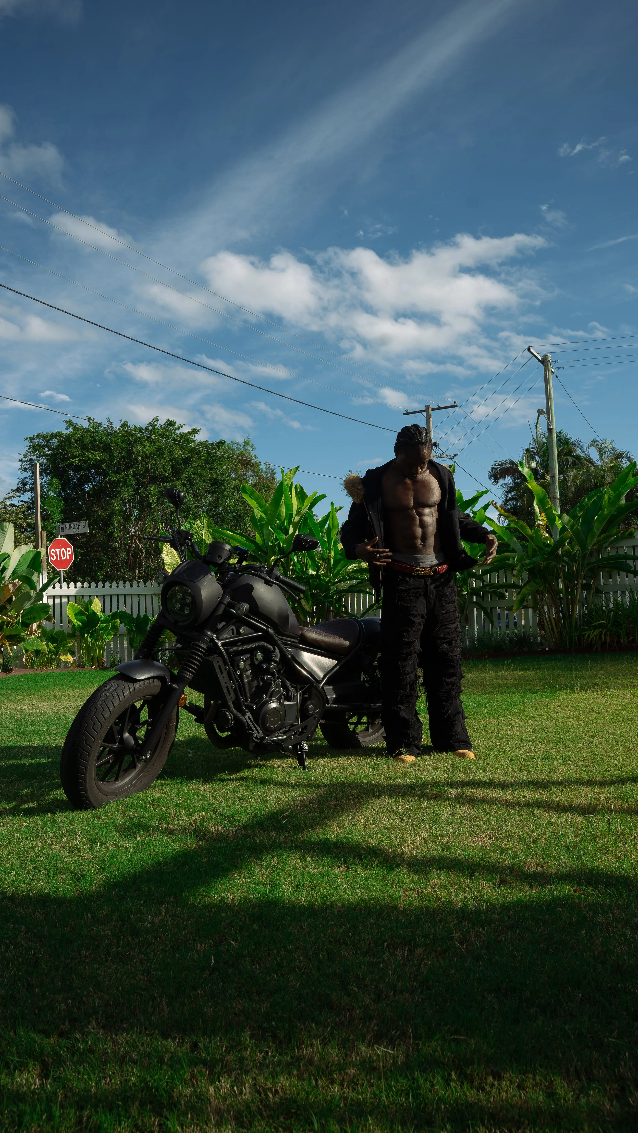 A shirtless man with a vest in his hand standing next to a black motorcycle on green grass, with a background of a white picket fence, tall green plants, and trees, under a partly cloudy blue sky.