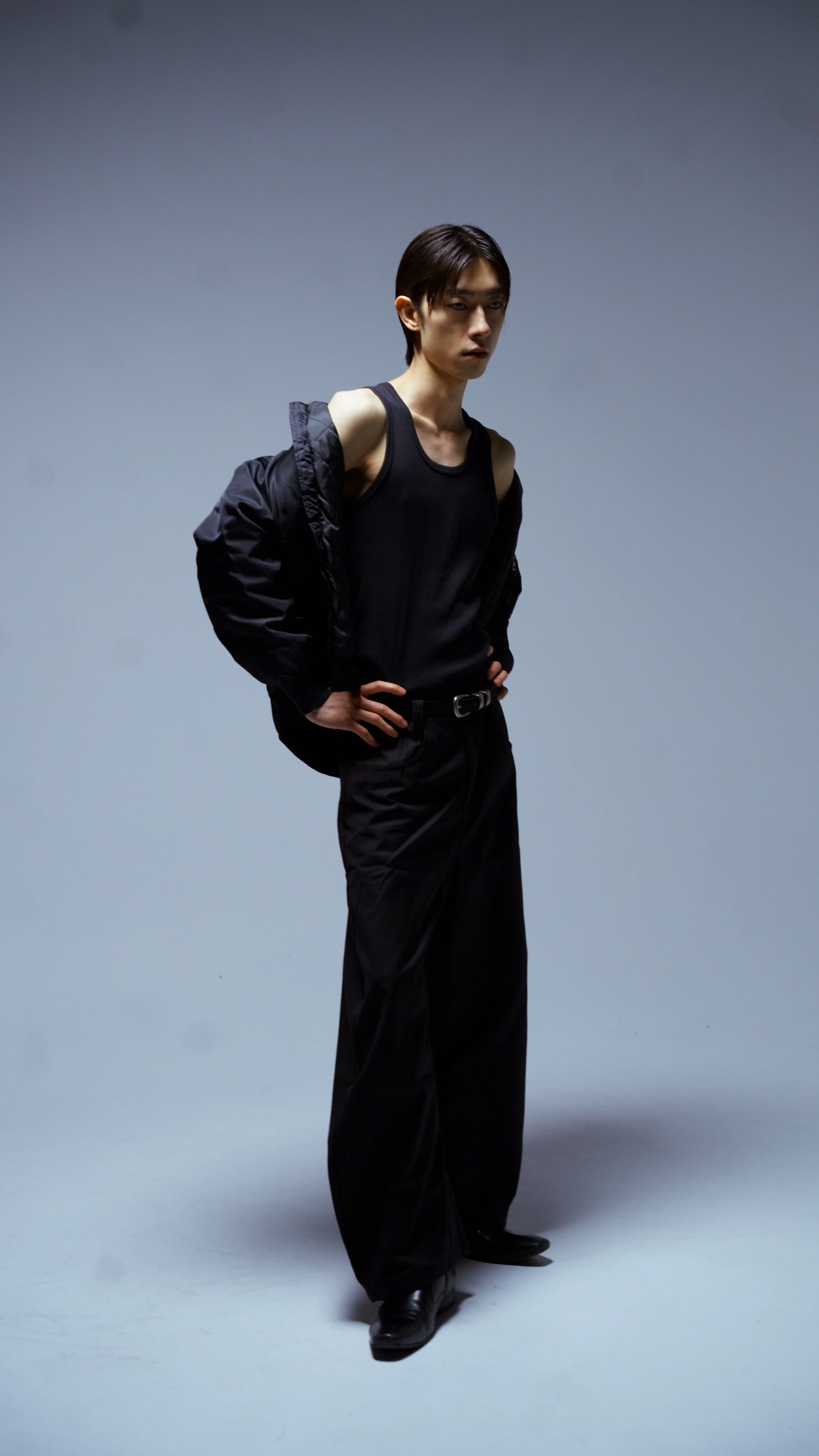 A young man with dark hair wearing a black tank top, black pants, black boots, and a black jacket draped over his shoulders, standing against a plain gray background.