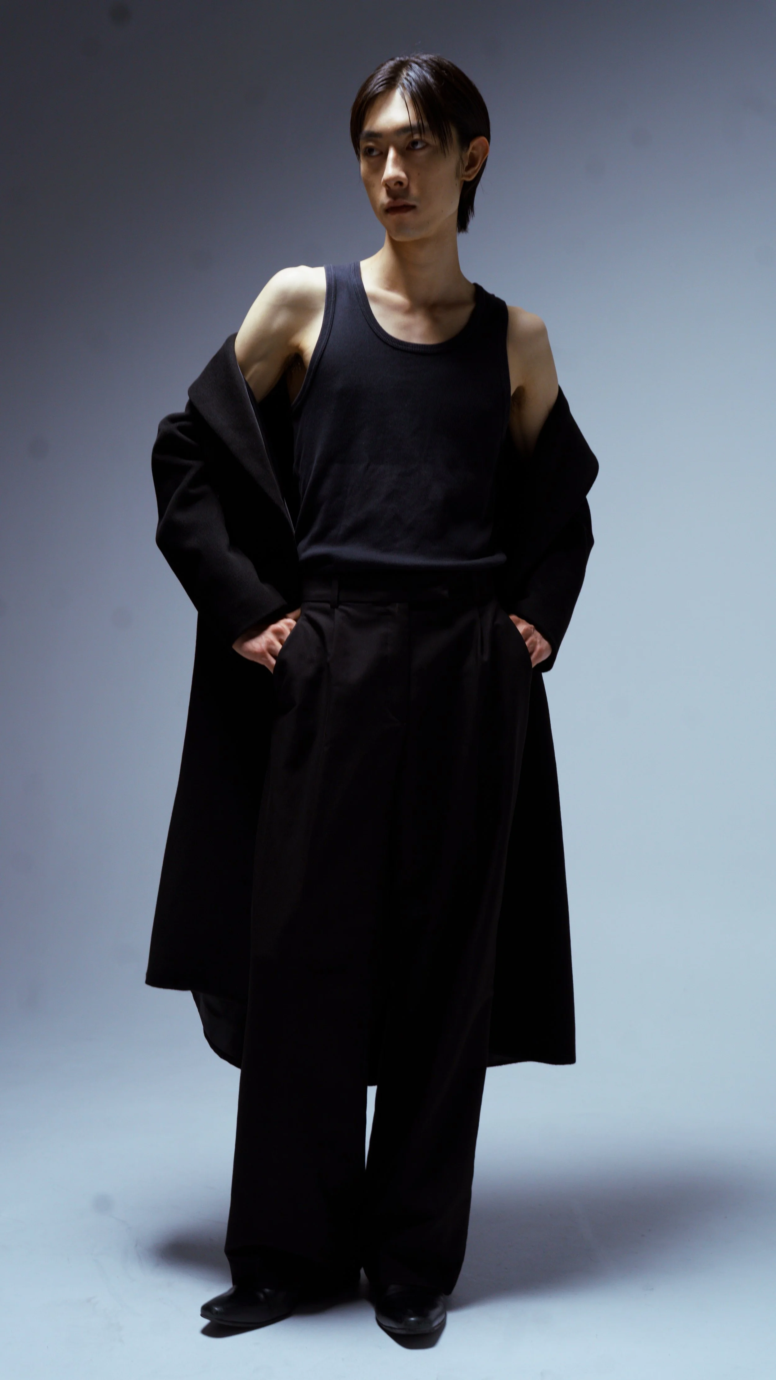 A person standing against a plain background, wearing a black sleeveless shirt, high-waisted black trousers, and a black coat draped over their shoulders, with hands in pockets.