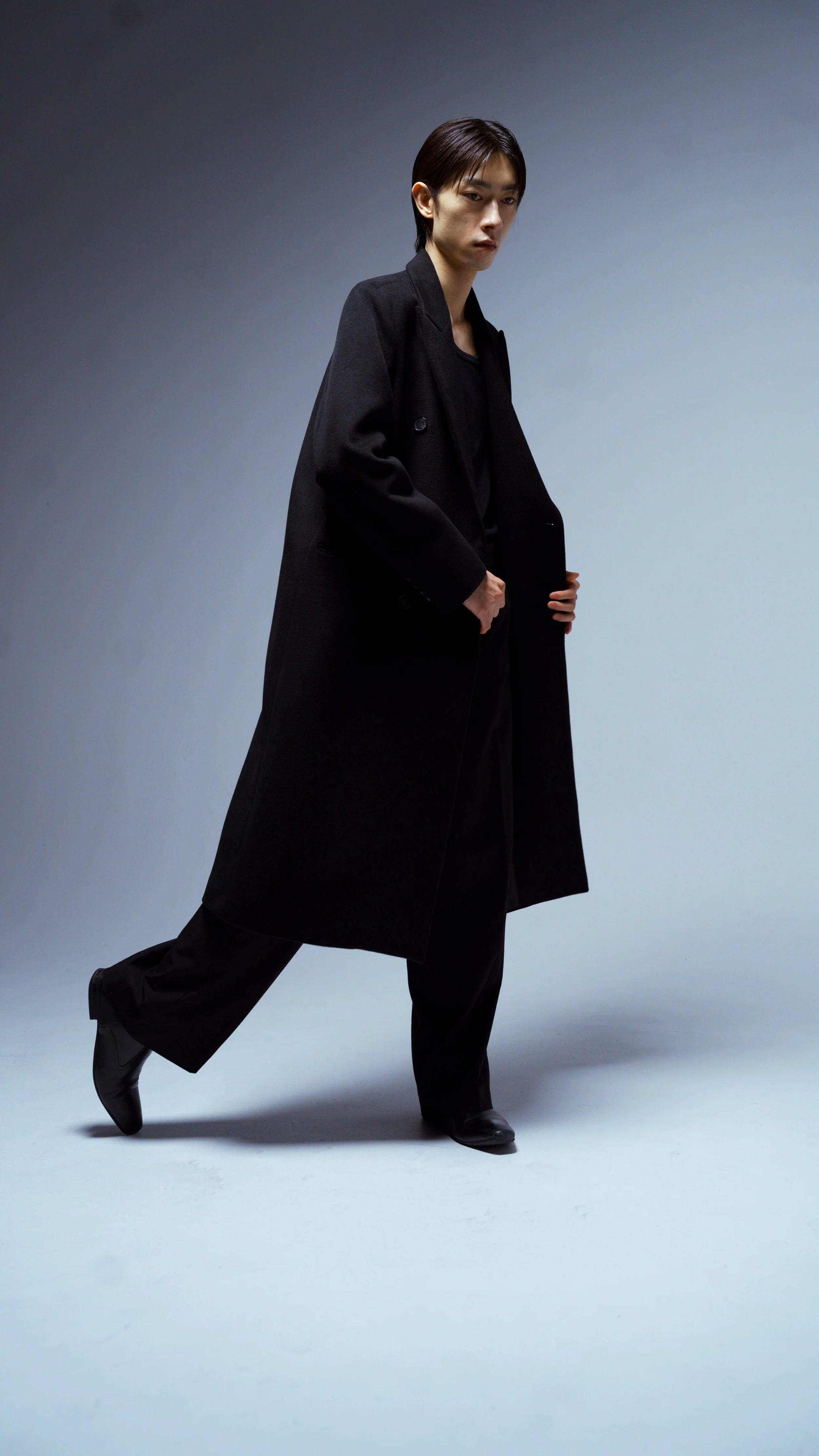 A person in a black outfit, including a long coat and dress shoes, standing against a gradient gray background.