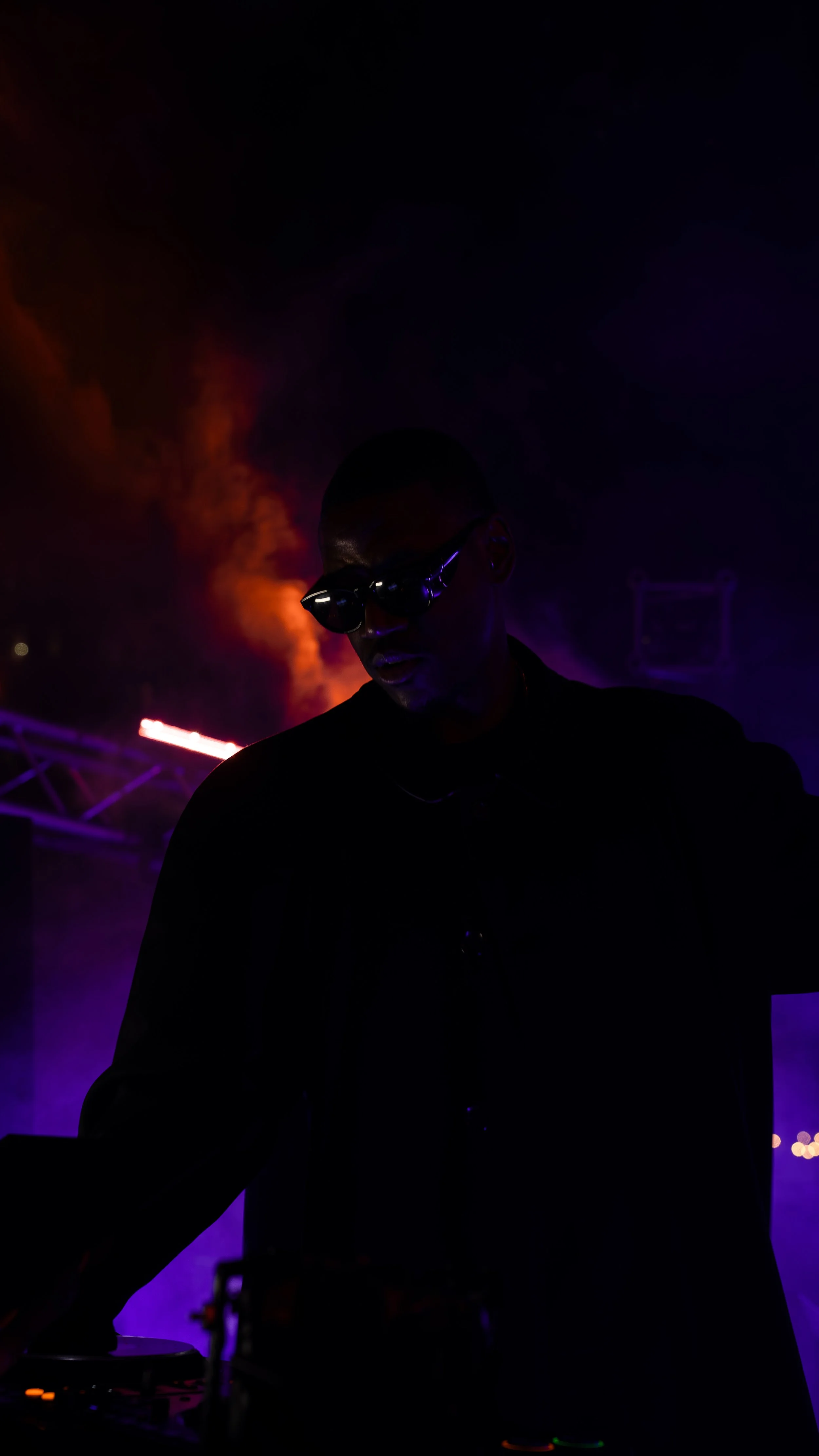 A silhouette of a DJ in a dark setting with colorful stage lights and smoke in the background.